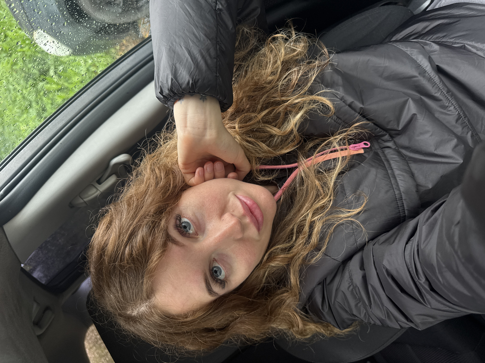 A young woman with wavy light brown hair and blue eyes is taking a selfie inside a car. She is wearing a black puffer jacket with a pink zipper. Her right hand is near her ear. Rain is visible on the car window. The location of the image cannot be confirmed in the city of Chaikovsky, Russia.