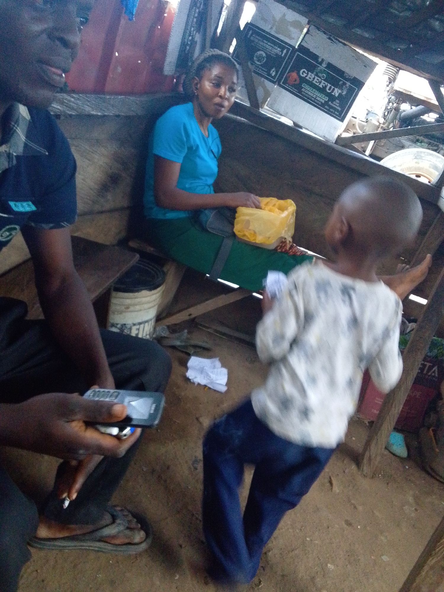 The image shows an informal indoor setting in Lokoja, Nigeria. Three people are visible: an adult male in the foreground holding a mobile phone and smoking a cigarette; a woman seated behind him with a yellow plastic bag; and a young child standing in the right of the frame. The indoor space appears to be a small, makeshift structure. A paint bucket and crumpled paper are on the floor. A box with the label "GBEFUN" is visible in the background, along with glimpses of vehicle parts.