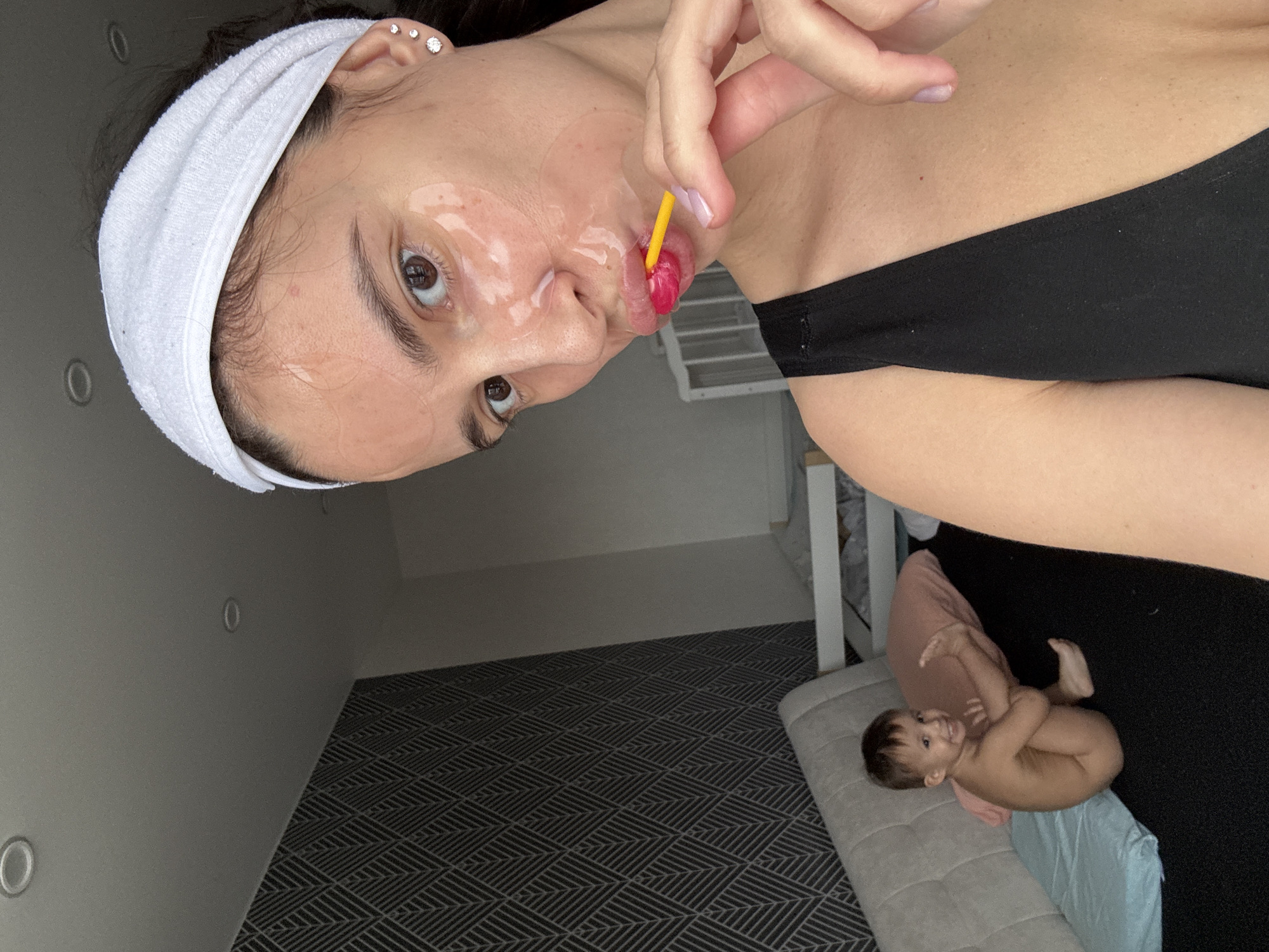 A woman with a white headband and black tank top is shown in a portrait-style photo, possibly a selfie. She has a transparent face mask on and is holding a lollipop with a yellow stick in her mouth. The setting appears to be a room with gray walls and a patterned carpet. In the lower right corner, a baby is lying on a gray bench with a light blue and pink fabric. The location is potentially Chaikovsky, Russia, based on the user's input.
