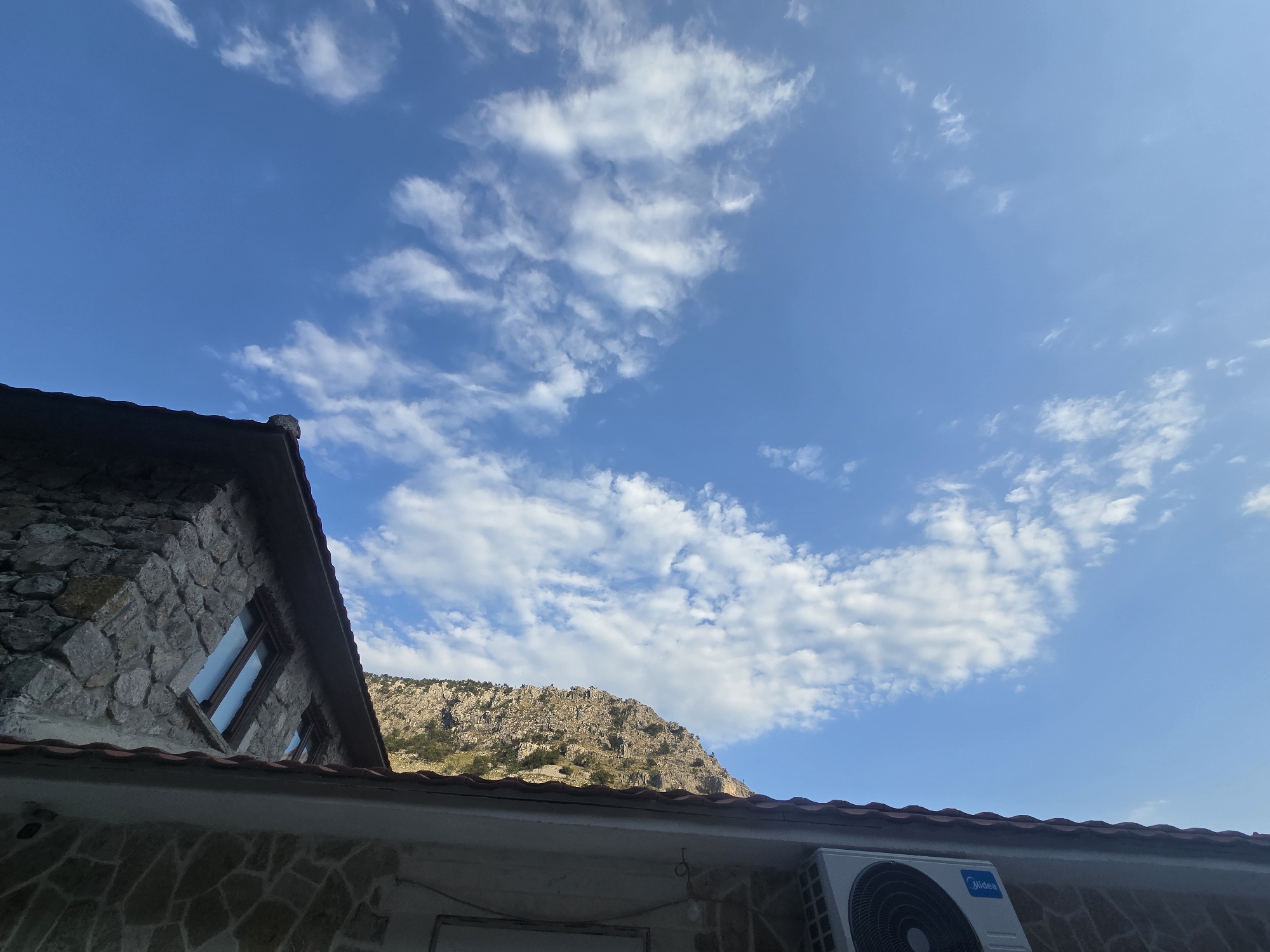 The image is an exterior, daytime, eye-level shot of a mountainous landscape with a partially visible building in the foreground. The sky is a bright blue with scattered white clouds. In the background, a rugged, rocky mountain range is visible, with some sparse vegetation on its slopes. In the foreground, the roofline of a building is prominent, with terracotta tiles. A portion of a stone-walled structure, likely part of the same building, is visible on the left, featuring a window. On the right side of the roof, an outdoor air conditioning unit is mounted. The image appears to be taken in Podgorica, Montenegro, given the characteristic mountainous terrain often surrounding the city. There are no people or animals present in the scene.