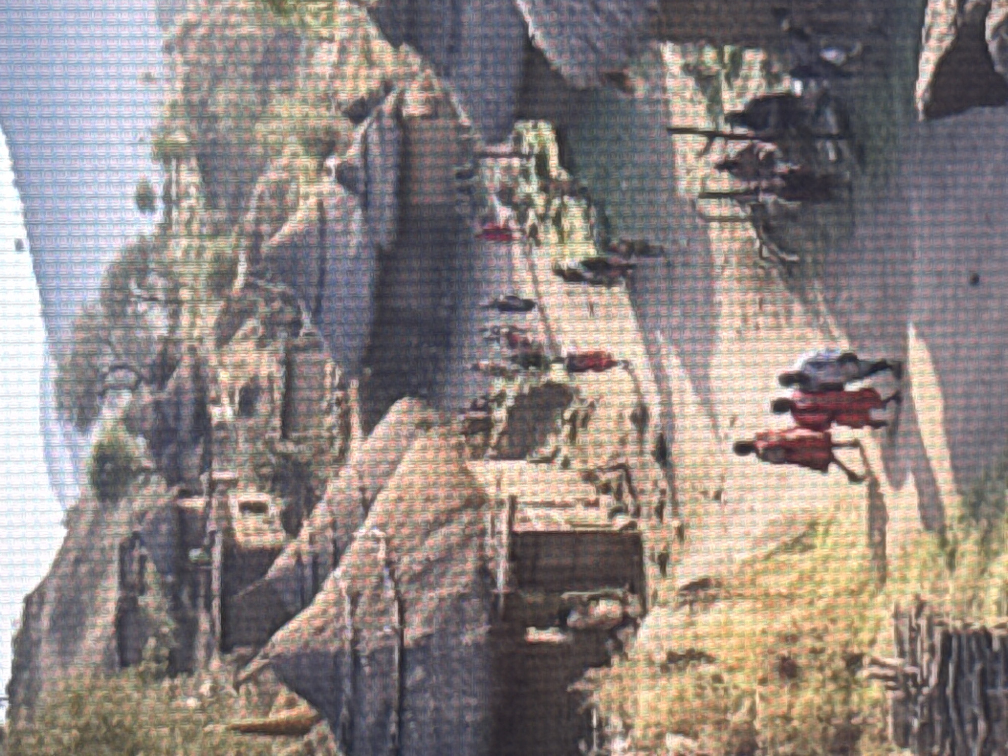 The image depicts a group of people in red clothing in an outdoor setting that appears to be a steep, rocky cliff face with sparse vegetation. The scene is rendered with a pixelated effect, suggesting it might be a screenshot from a video game or a low-resolution digital image.
The subjects are several individuals, each wearing red attire. Some appear to be in motion, with limbs extended, suggesting they are engaged in an activity. There are also objects visible in the scene, including what look like structures built into the cliff face, possibly dwellings or platforms. Further down the cliff, there are more figures and what might be pathways or stairs.
The setting is a dramatic, vertical landscape dominated by large rock formations. The vegetation is green and appears to be growing in patches on the rocks and at the base of the cliff. The lighting suggests it is daytime, with shadows cast by the terrain and the figures. The overall impression is one of an adventurous or possibly dangerous activity occurring in a challenging environment. The pixelation makes it difficult to discern specific details like facial expressions or the precise nature of the structures. There is no visible text in the image.
