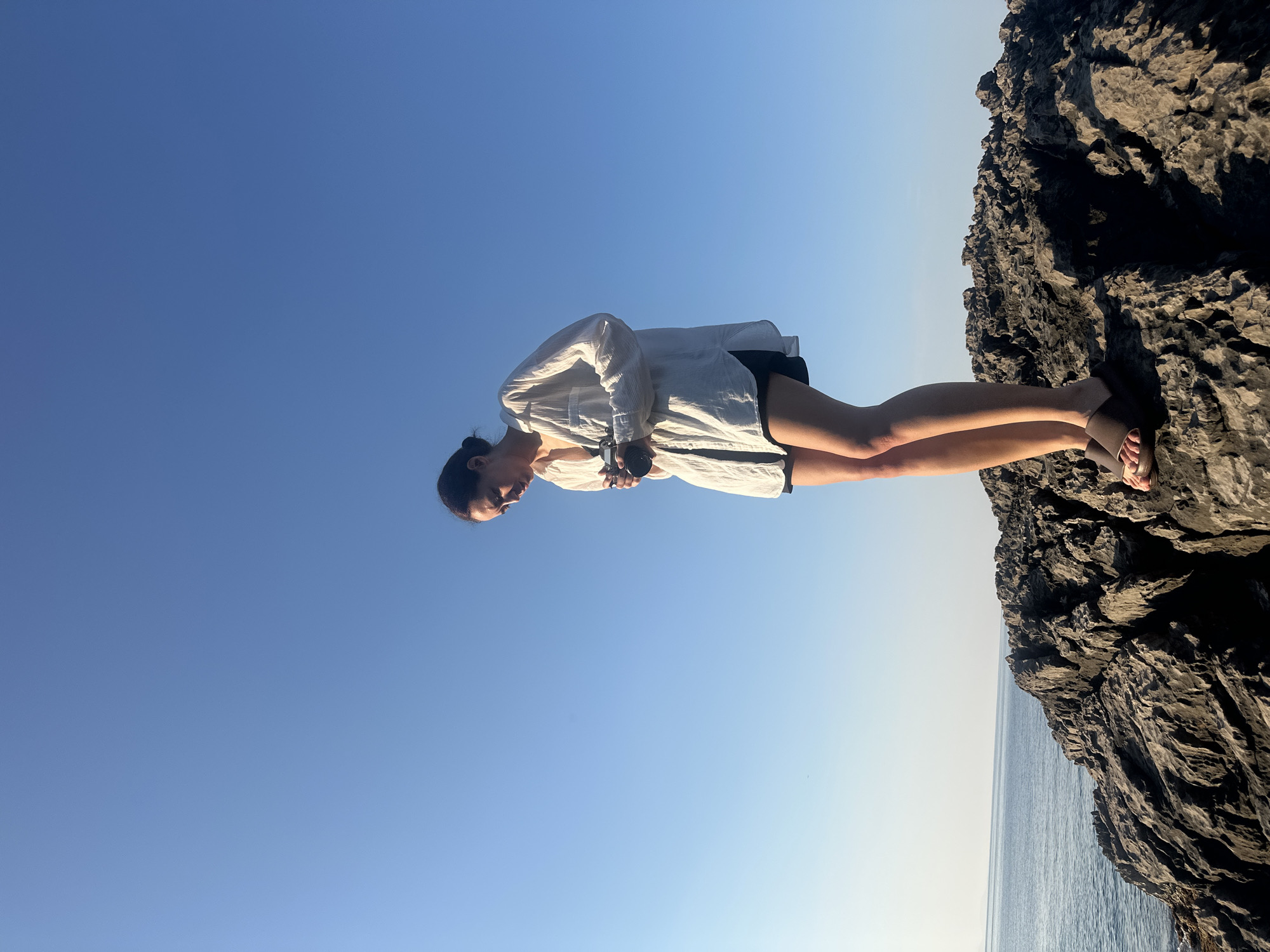 A young woman is captured in a vertical, low-angle shot, standing on a rocky outcrop with the vast expanse of a clear blue sky behind her. She is wearing a white, loose-fitting button-down shirt over dark shorts, and a pair of sandals. Her hair is pulled back in a bun, and she is looking downwards, holding a dark, cylindrical object in her hands, possibly a camera or a phone. Her posture suggests a moment of contemplation or focus.
The scene is set by the sea, with the calm, blue water visible in the lower right portion of the image. The rocks themselves are rough and textured, indicating a natural, coastal environment. The bright, clear sky suggests it is daytime, likely late afternoon or early morning due to the quality of light illuminating the woman's face and casting subtle shadows on the rocks. The overall mood is serene and tranquil, with a sense of connection to nature. There is no visible text in the image.