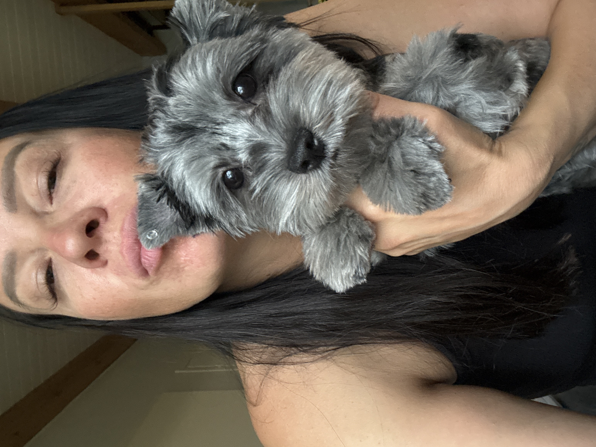 A woman holds a small grey dog in her arms, looking towards the camera with her lips pursed. The woman has long, dark hair and is wearing a black tank top. The dog has bright eyes and fluffy fur. The background appears to be an indoor setting with wooden beams visible on the ceiling, suggesting a possibly rustic or attic-like environment. There is no indication of the city of Chaikovsky, Russia, in the image or its context.