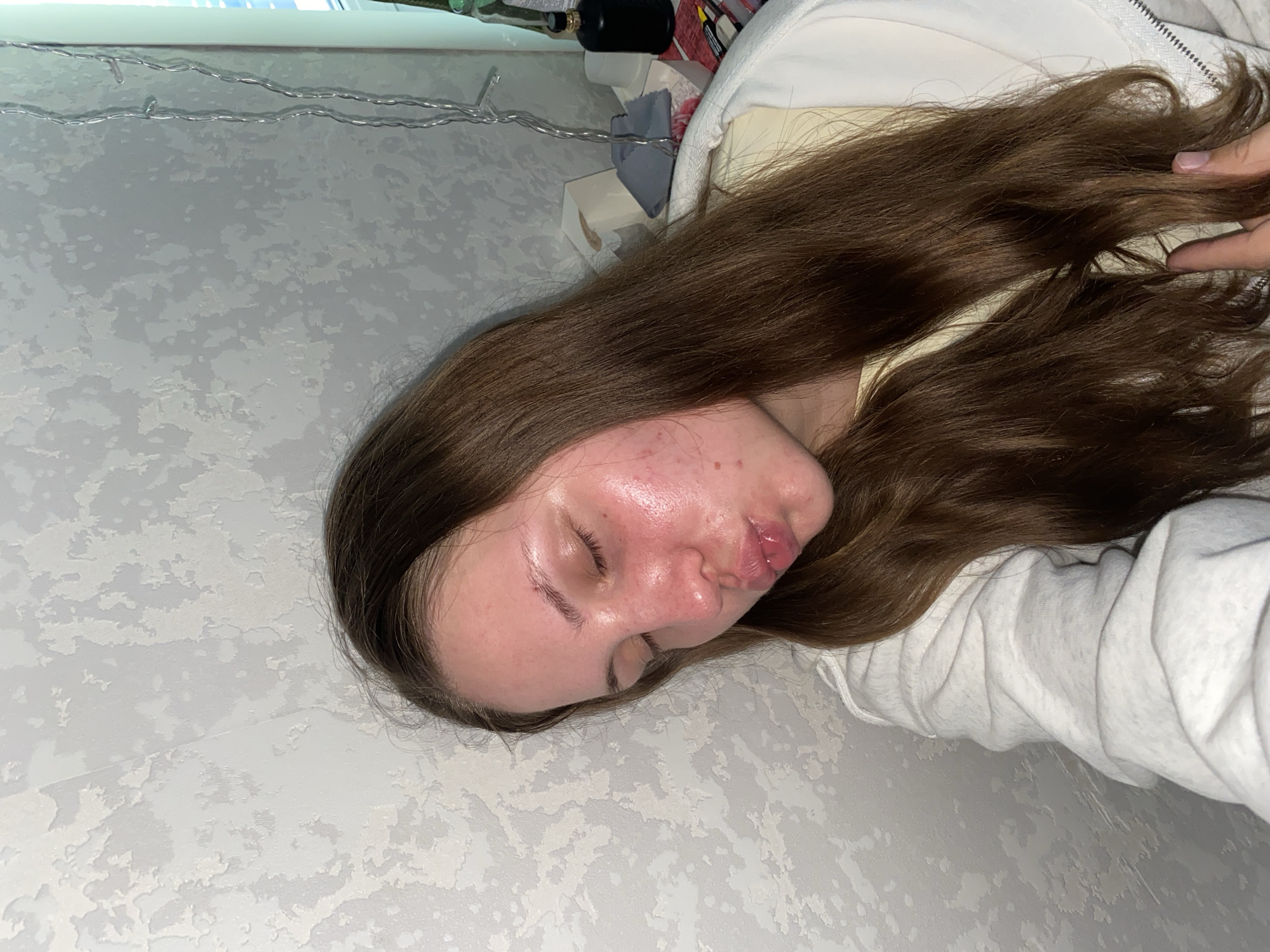A young woman with long brown hair, wearing a white hooded sweatshirt over a yellow garment, is captured in a close-up selfie. Her eyes are closed, and her lips are pursed in a kissing gesture. Her skin appears flushed and has visible blemishes. She is indoors, with a textured, light-colored wall as the backdrop. A string of fairy lights hangs vertically on the right side of the frame, and in the background, several objects are faintly visible, including what appears to be tissue boxes, a gray cloth, and a dark bottle. The image does not provide any specific clues to its location within the city of Izhevsk, Russia, or any identifiable landmarks.