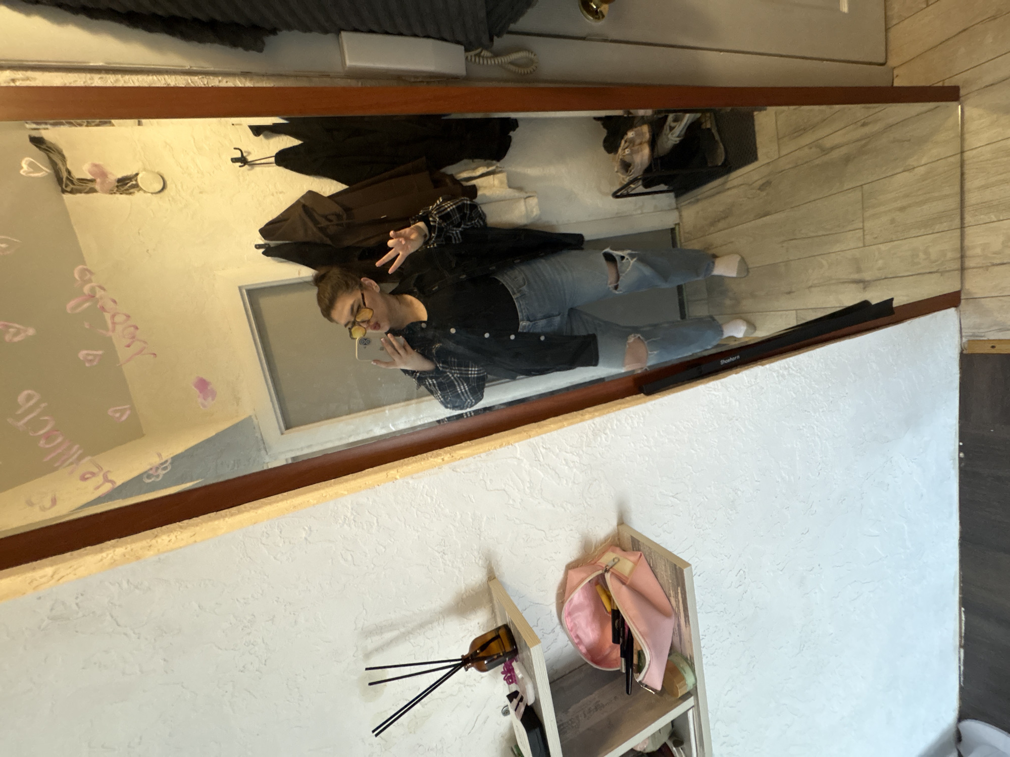 The image shows a mirror reflecting a young woman in ripped jeans and a plaid shirt taking a selfie. She is wearing glasses and making a peace sign. The reflection also shows part of a room with a closet, a shelf with cosmetic items, and a wall. The location is in Chaikovsky, Russia. The writing in the mirror on the top-left translates to "tenderness."