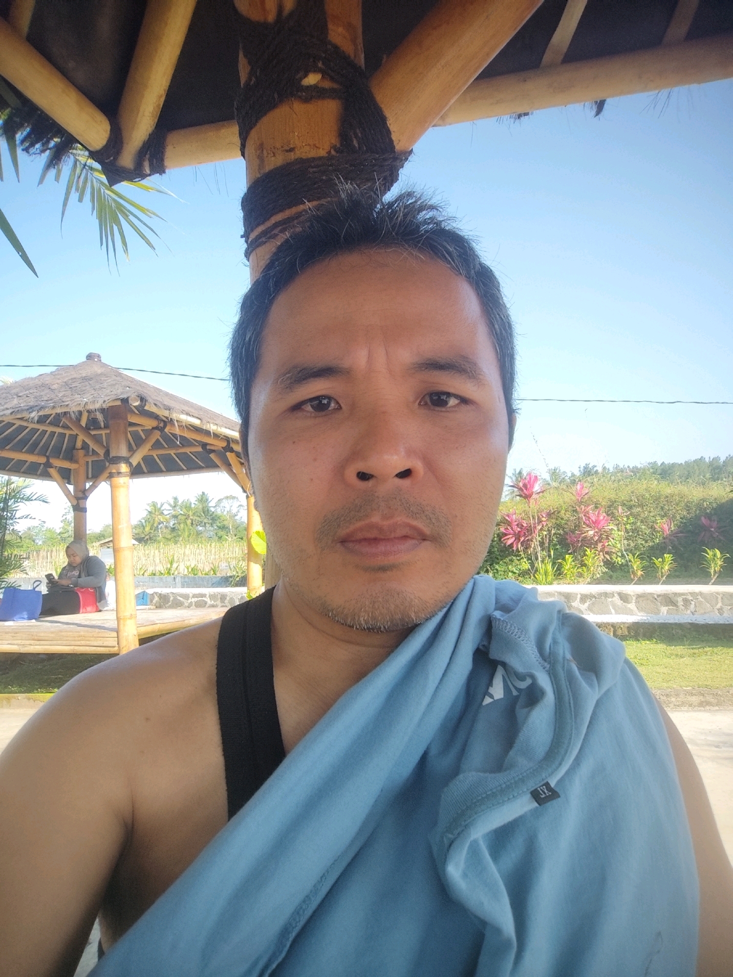An outdoor, medium shot selfie shows a man in the foreground, facing the camera. He is wearing a black tank top, and a light blue t-shirt is draped over his right shoulder and chest. His hair is dark and slightly disheveled. The background features a thatched-roof gazebo with bamboo supports, and a person wearing a hijab is sitting inside it, looking at a phone. Beyond the gazebo, there are lush green trees, flowering plants with pink and green leaves, and a clear blue sky. The setting appears to be a tropical outdoor recreational area.