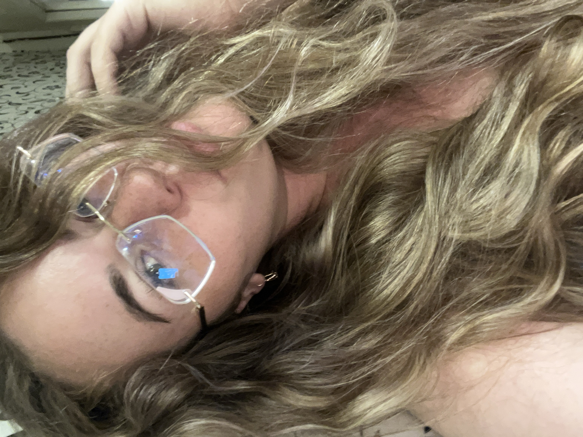 A young woman with blonde, wavy hair and glasses is looking at the camera, touching her hair with her right hand. She is wearing a small hoop earring in her visible left ear. The background appears to be an indoor setting with a patterned wallpaper and a hint of a wall on the right. The image does not provide any information about the location in the city of Chaikovsky, Russia.