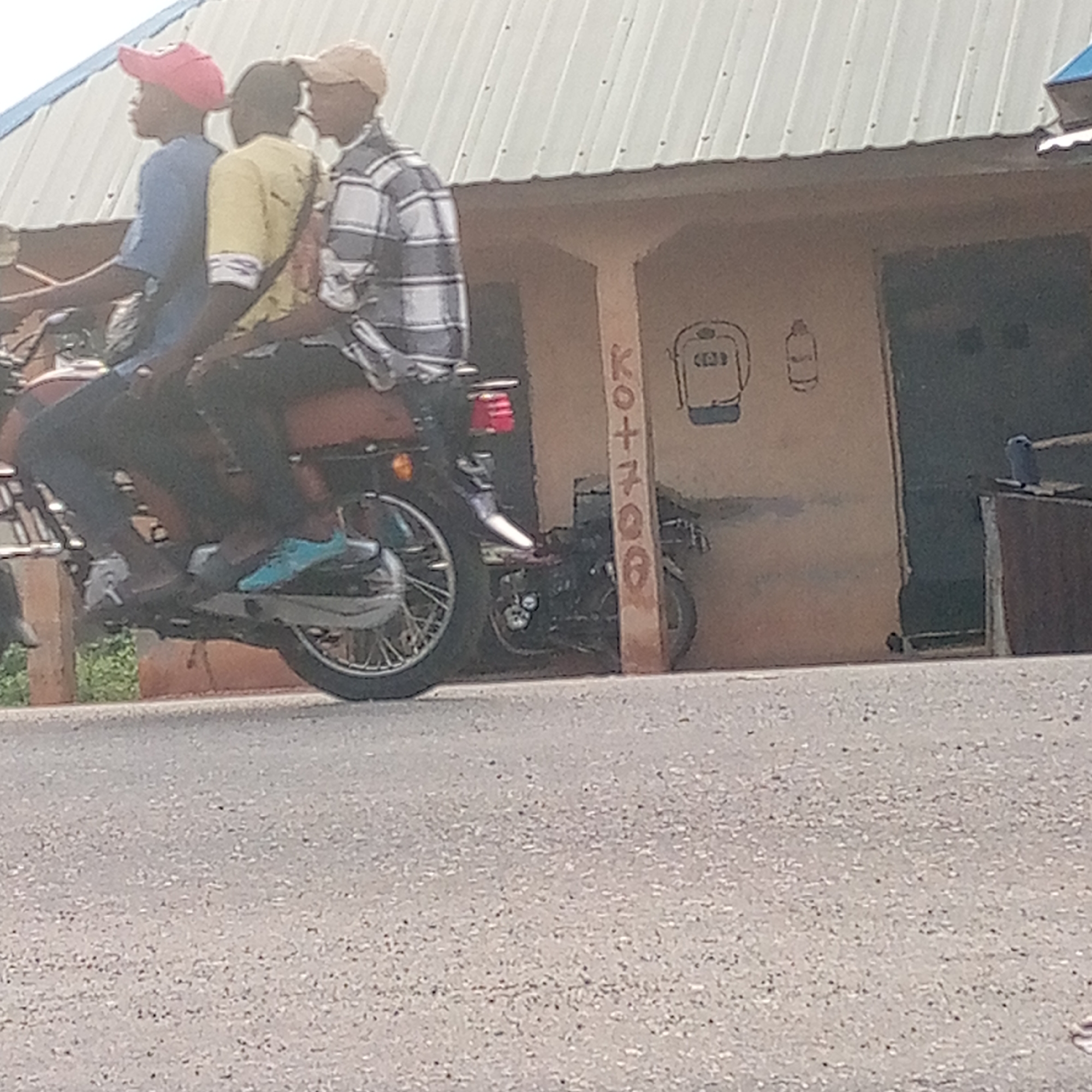 The image shows three men riding a single motorcycle on a paved road. The first man is wearing a red cap and blue shirt. The second man is wearing a yellow shirt, and the third is wearing a plaid shirt and tan cap. The motorcycle is black and red. The background shows a building with a tin roof. The building has a painted advertisement on the side, which appears to say "KO+7008" along with a drawing of a bag. Another motorcycle is parked in front of the building. The scene is set in Mararaba, Nigeria, as indicated in the prompt.