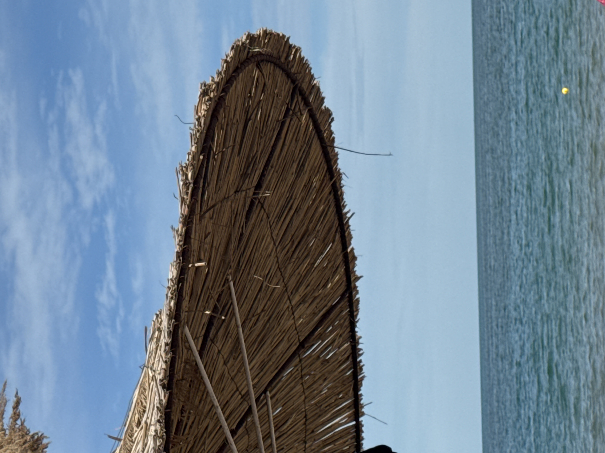 This is a close-up, eye-level, vertical, outdoor, full shot of a thatched beach umbrella casting a shadow over a beach. The umbrella is positioned in the foreground, taking up the left side of the frame. The umbrella's texture is rough, made of straw or reeds, and appears dry and weathered. The sky above is a clear blue with scattered white clouds. Below the umbrella is the ocean, a vast expanse of blue water with gentle waves. The horizon line is visible where the sea meets the sky. There is a small yellow buoy in the water. This scene is likely from a beach in Varna, Bulgaria, a city known for its coastline and resorts. No people are clearly visible in the frame. The overall impression is one of a sunny, peaceful day at the beach.