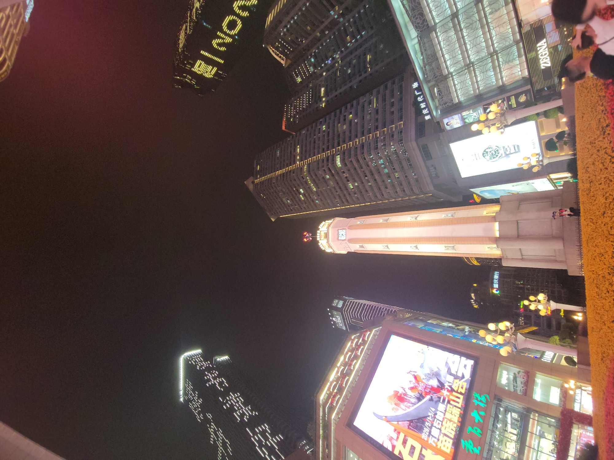 The image presents a dramatic low-angle view of a vibrant city center at night, dominated by illuminated skyscrapers and urban infrastructure. The dark night sky contrasts sharply with the numerous artificial lights emanating from buildings, signs, and streetlights.
Several tall buildings define the skyline, glowing with light from windows and exterior architectural features. One building on the left edge features a vertically oriented, illuminated sign that appears to read "LUXONE." Another prominent building on the right clearly displays the brand name "ZEGNA." A central, light-colored tower with a classical design stands out, topped by a visible clock face.
In the lower-right foreground, a large digital billboard is a focal point, displaying an advertisement with an animated scene of figures in traditional attire, possibly depicting a cultural festival. Overlaying text on the billboard includes "2026/5/1-5/4" and Chinese characters "万魔石雄第二十六 苗族踩山含云," which translates to a "Miao Ethnic Cai Shan Han Yun" festival, indicating a future event. Below this, a sign reads "重庆时代广场" (Chongqing Times Square).
To the far right, a decorative barrier covered in yellow and red flowers or foliage runs vertically, with several people visible beyond it, observing the bustling urban environment. Decorative streetlights with multiple glowing globes are positioned throughout the lower and mid-sections of the frame. Other discernible text on various buildings includes "重庆银行" (Chongqing Bank) and "委万大楼" (Weiwanda Building). The scene captures the lively and impressive atmosphere of a modern metropolis, consistent with Yuzhong District, Chongqing, China.