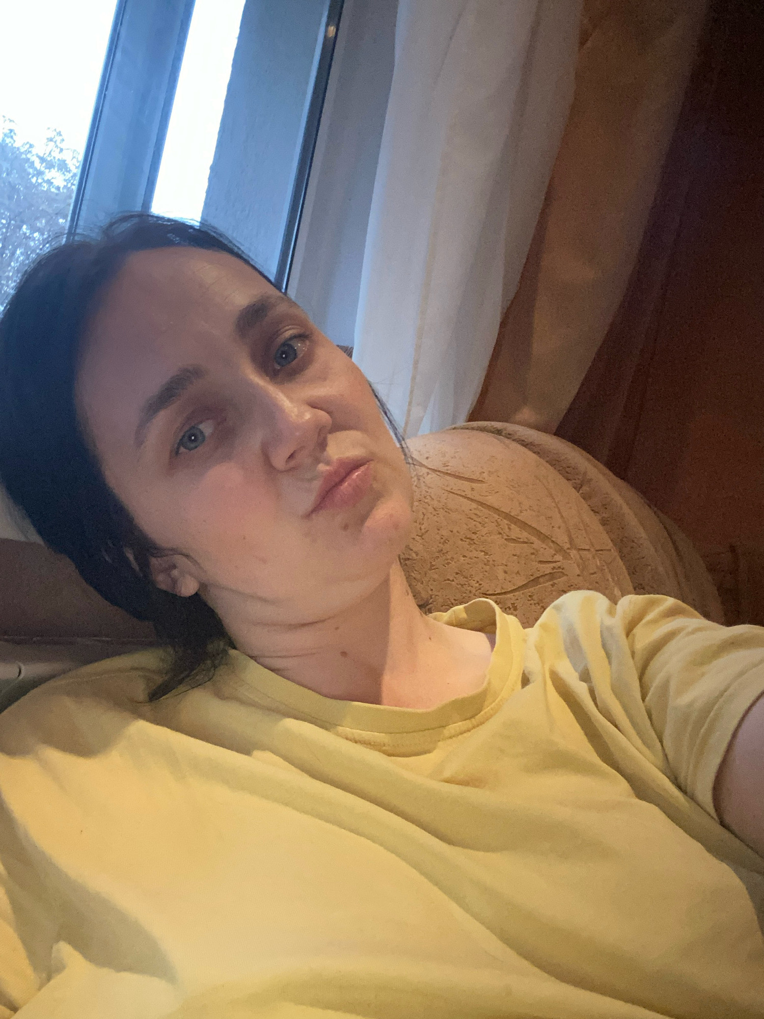 A woman is captured in a selfie, leaning back with her head tilted. She has dark hair and blue eyes, and she is wearing a yellow t-shirt. Her lips are pursed, and her gaze is directed towards the camera. The background shows a window with a view of trees outside, and sheer curtains. The woman appears to be indoors, possibly in a living room or bedroom, with a plush, textured piece of furniture behind her. The lighting is soft, suggesting it might be daytime or evening.