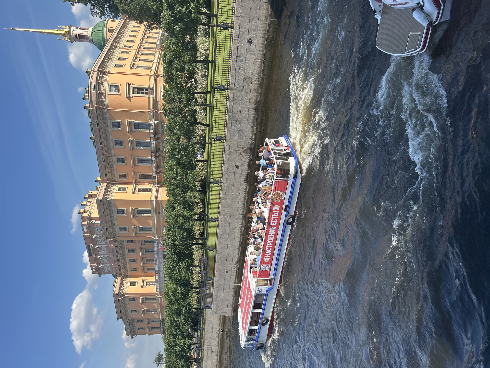 This is a full shot of the Mikhailovsky Castle in Saint Petersburg, Russia, on a bright sunny day. The castle is a large, salmon-colored building with many windows, topped by a green dome and a tall golden spire. In front of the castle, a treeline runs along a stone embankment. A tour boat filled with people is moving down a dark blue river in the foreground. The boat is white with red trim and has advertisements on its side. Another smaller boat is visible to the right, partially submerged in the water and creating a wake. The sky is clear with a few scattered white clouds.