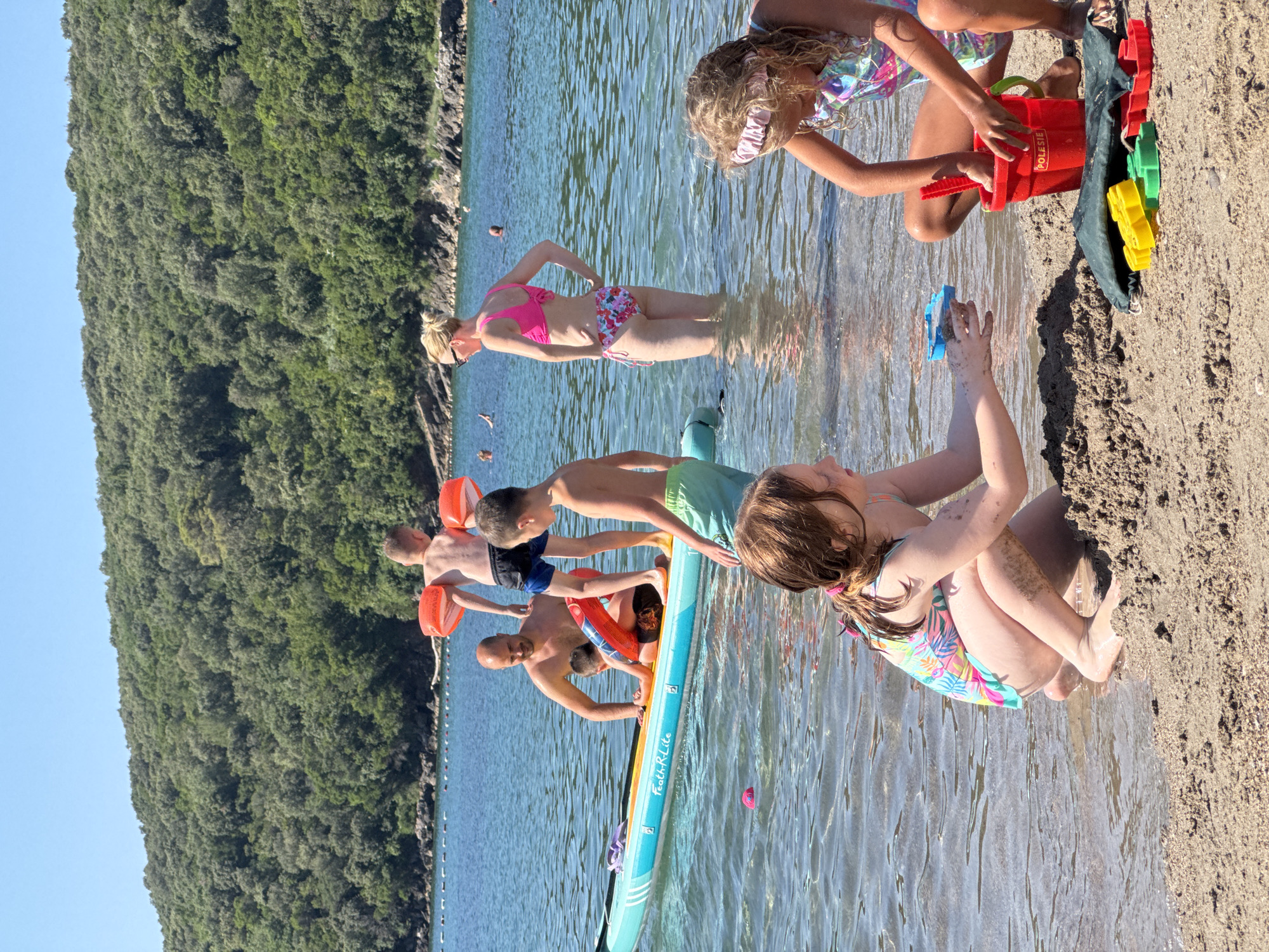 The image depicts a group of people at a beach in Prijevor, Montenegro. The scene is set on a sunny day with clear blue water and a lush, green forested hillside in the background. Several individuals, including adults and children, are interacting in and around the shallow water.
In the foreground, a child with blonde hair is partially submerged in the water, reaching for a blue toy. Next to them, a woman in a pink bikini stands in the water, her hand on her hip. A group of boys are gathered on a stand-up paddleboard, some holding bright orange flotation devices. One boy is reaching towards a red bucket filled with sand toys. Further back, another child is sitting on the sandy shore, also playing with sand and toys.
The overall mood appears to be relaxed and joyful, indicative of a family outing or a day at the beach. The water is calm and reflects the sunlight, creating a shimmering effect. Visible text on the paddleboard reads "Feath-R-Lite". The presence of sand toys suggests children are engaged in typical beach activities. The time of day is likely midday, given the bright sunlight and shadows.