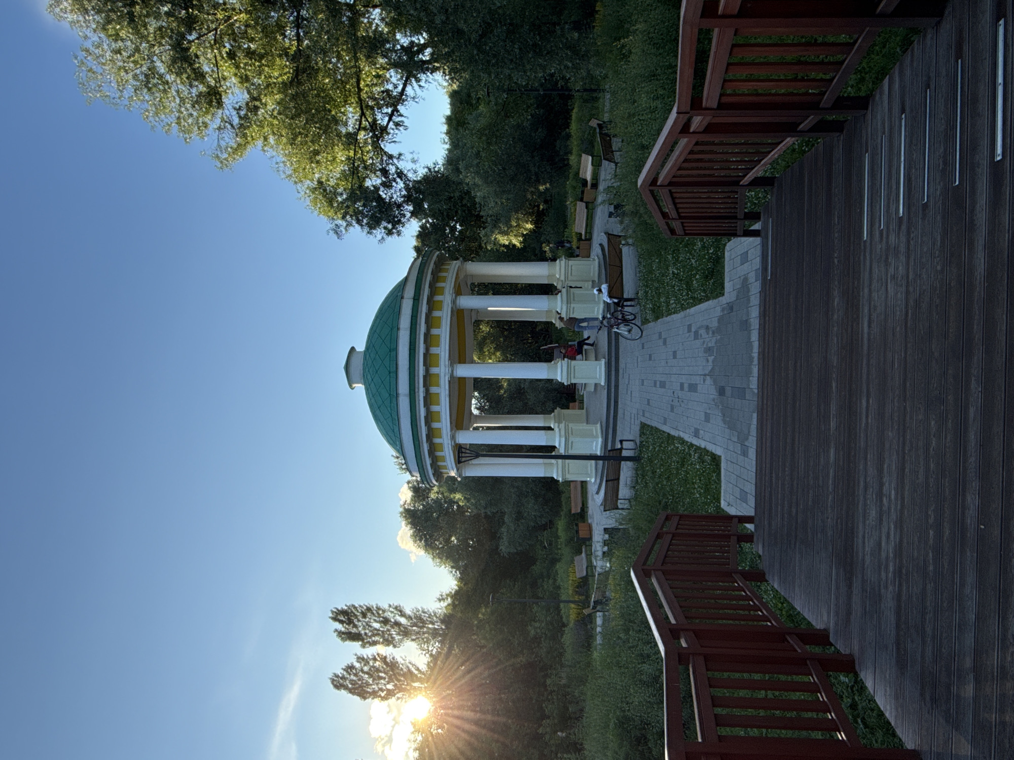 The image depicts a gazebo with a green dome and white pillars, situated in a park. A paved pathway leads to the gazebo, and there are benches visible along the path. The setting appears to be a serene park with lush green trees and grass, suggesting a pleasant outdoor environment. To the right of the gazebo, there is a wooden deck with railings, adding a modern touch to the natural setting.
In the foreground, a bicycle is leaning against the gazebo, and a few people are seated inside or near the structure. The time of day appears to be late afternoon or early evening, indicated by the low angle of the sun casting a warm glow and creating sunbursts. The sky is clear and blue, suggesting good weather.
The presence of the gazebo, pathways, benches, and trees points to a public park designed for relaxation and recreation. The wooden deck might be part of a picnic area or a viewing platform. The overall emotion conveyed by the scene is one of tranquility and natural beauty. There is no visible text in the image. Given the location context of Moscow, Russia, this scene could be set in one of its many parks, offering a peaceful escape from urban life.
