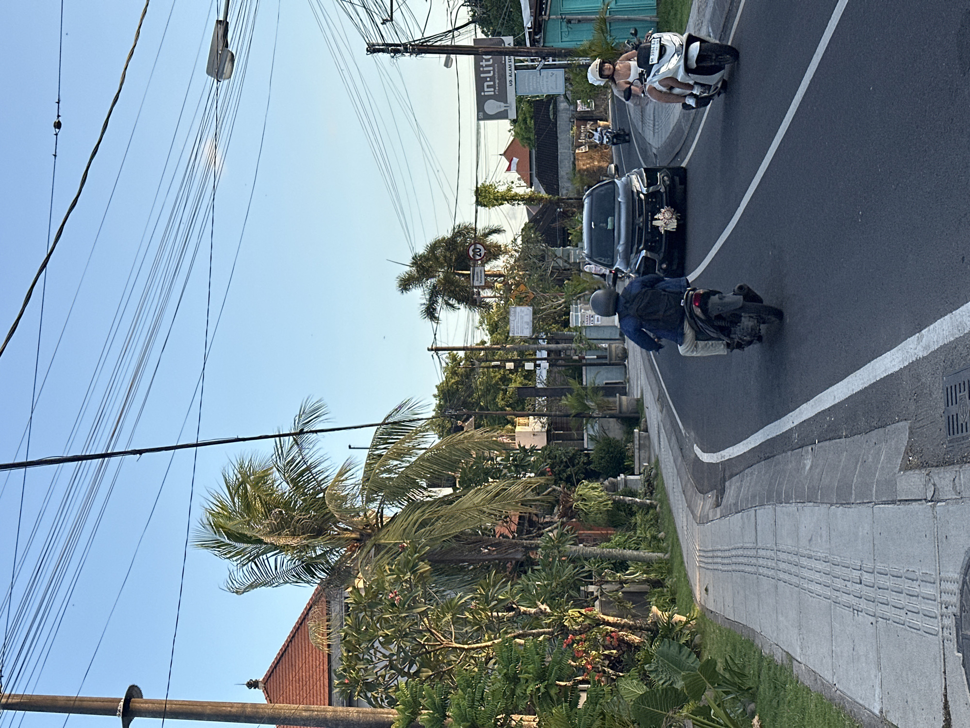 The scene is a street in North Kuta, Indonesia, with power lines visible against a clear blue sky. The road curves and has a white line dividing the lanes. A black car, possibly part of a wedding procession based on the decorations on its hood, is driving on the right side of the road. A man on a motorcycle is following behind the car, while a woman on a scooter wearing a white helmet is overtaking the car. The street is lined with sidewalks, trees, and buildings. There's a sign visible with "in-lite" written on it. Palm trees and tropical vegetation are also present.
