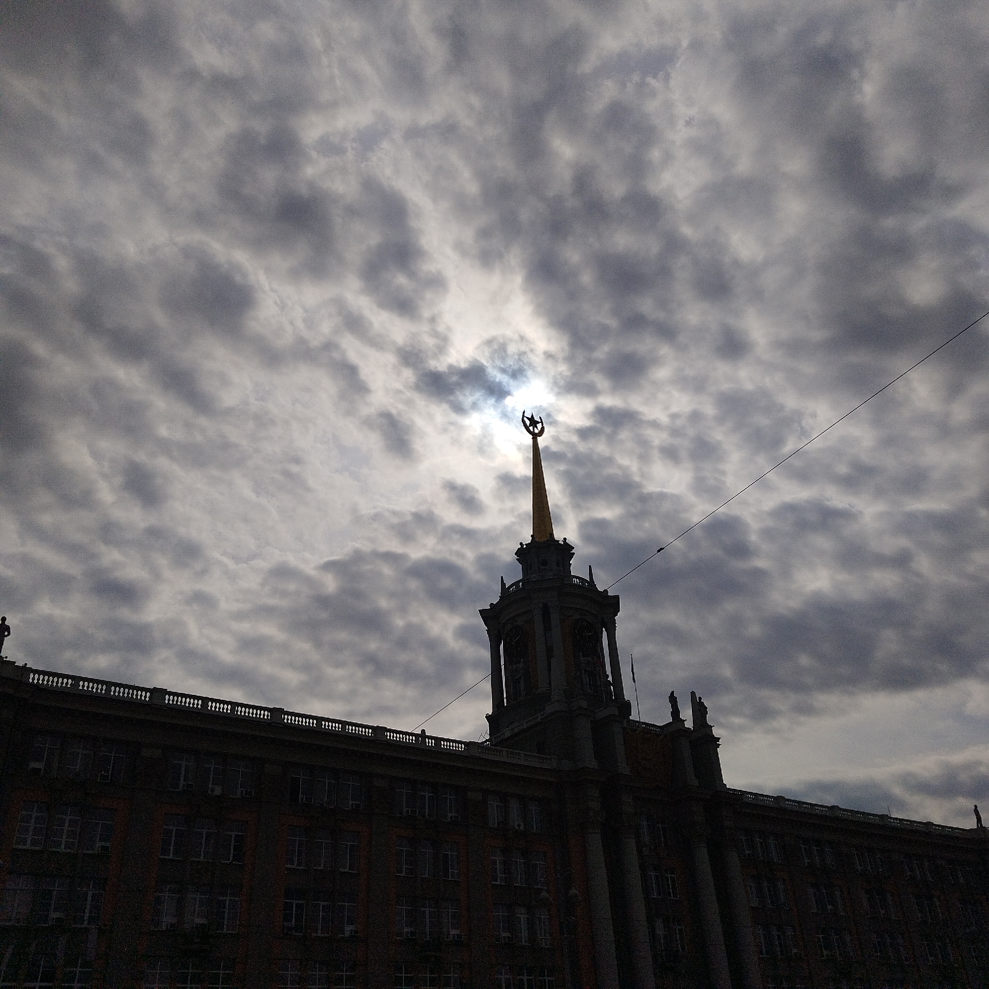 The image shows a low-angle view of a large building in Yekaterinburg, Russia, silhouetted against a cloudy sky. The building's architectural style suggests a Soviet-era construction, with a prominent central tower topped by a spire and a crescent moon symbol. The tower is positioned directly in front of a break in the clouds, with the sun shining through. The sky is filled with dark, textured clouds. A thin wire is visible across the upper right of the frame. The building's details, such as windows, columns, and rooftop statues, are partially obscured by the overall darkness. The base of the building is covered in greenery.