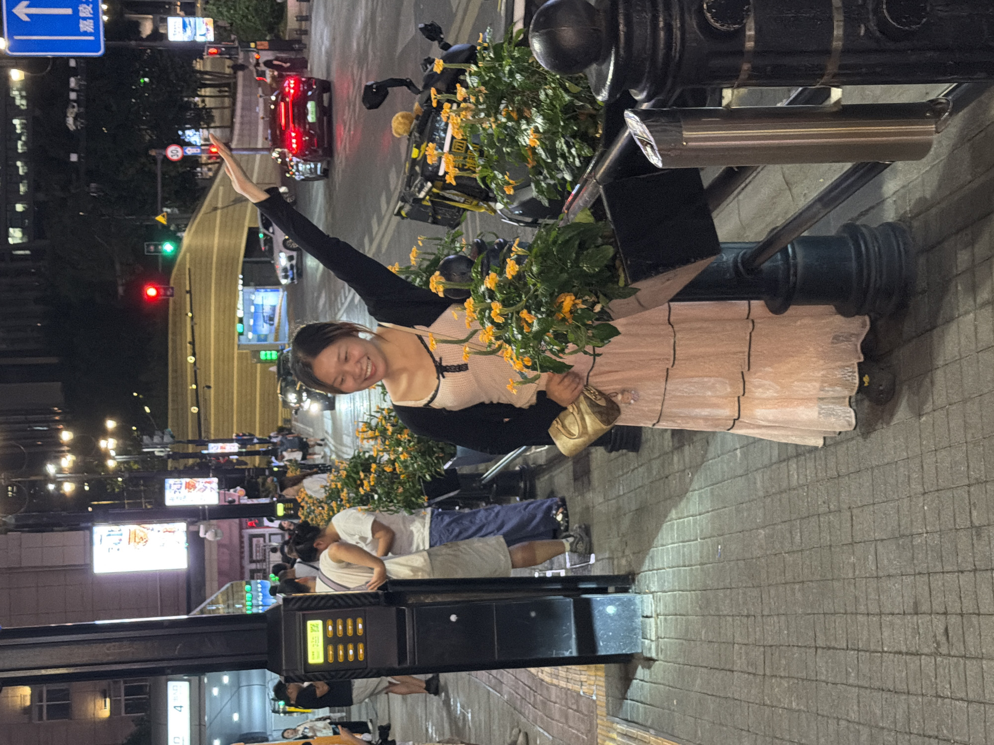 A young woman with brown hair is smiling and waving with her right hand while holding a small, light-colored purse in her left. She is wearing a light pink, knee-length dress with a black cardigan over a cream-colored top with black trim. The scene appears to be a city street at night, possibly in Yuzhong District, China, given the location context. The woman is standing near a metal railing with decorative black elements, and there are flowering plants with yellow blossoms in planters.
In the background, a red car is stopped at a traffic light. Several streetlights illuminate the area, casting a warm glow on the wet pavement. People are visible in the background, some walking and others appearing to be sitting or standing. A large screen displaying an image of food is visible on a building to the left. A blue street sign with white Chinese characters and an arrow is also present in the upper left corner of the image. The overall atmosphere is lively, suggesting a bustling urban environment in the evening. The wet pavement suggests it may have recently rained or is raining lightly.