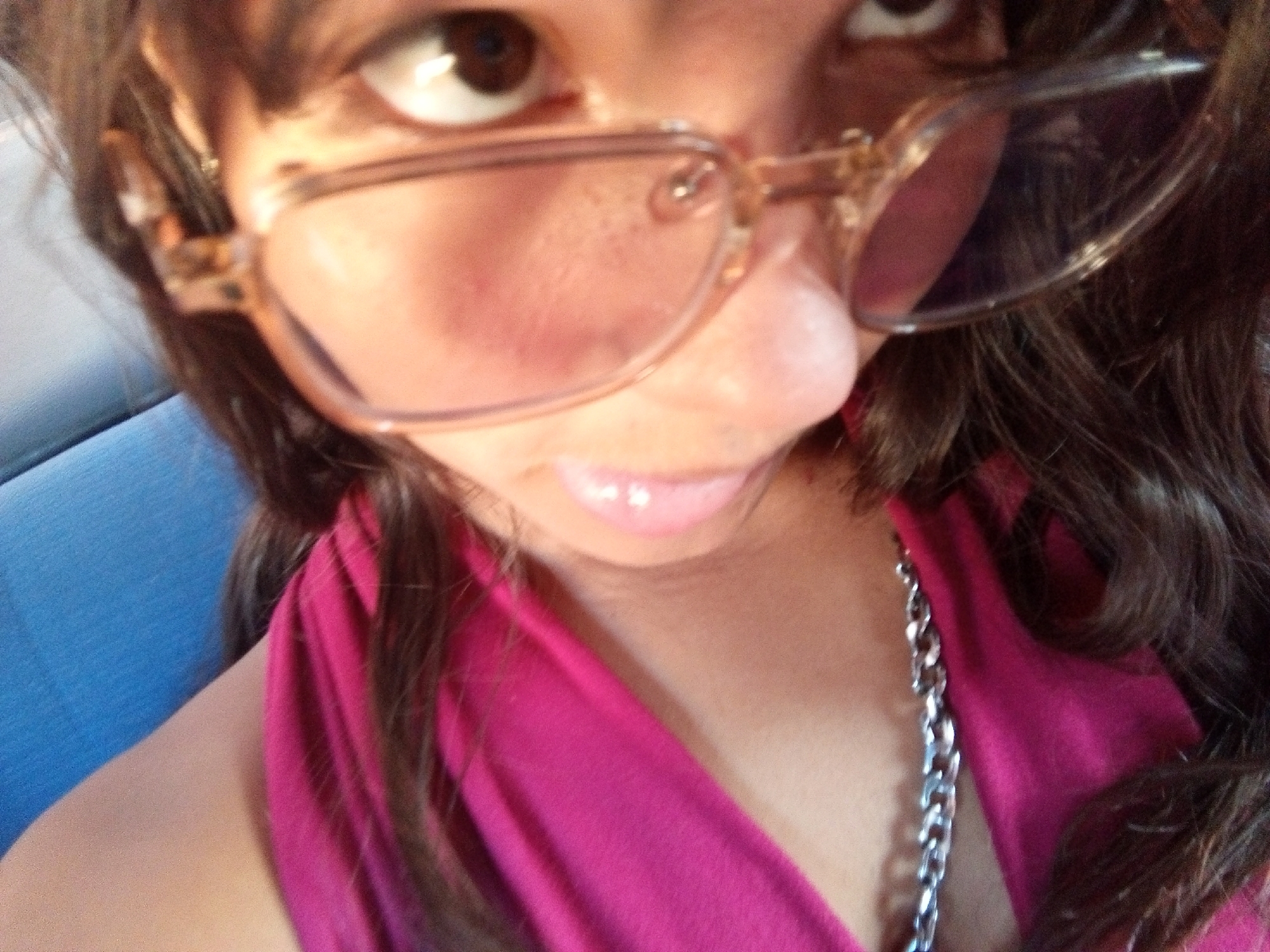 A close-up selfie of a woman with brown hair and brown eyes wearing tan-framed eyeglasses. She is wearing a bright pink top and a silver chain necklace. The location is possibly inside a vehicle with a blue seat. There is no definitive indication of the city of Juan Griego, Venezuela, in the image.