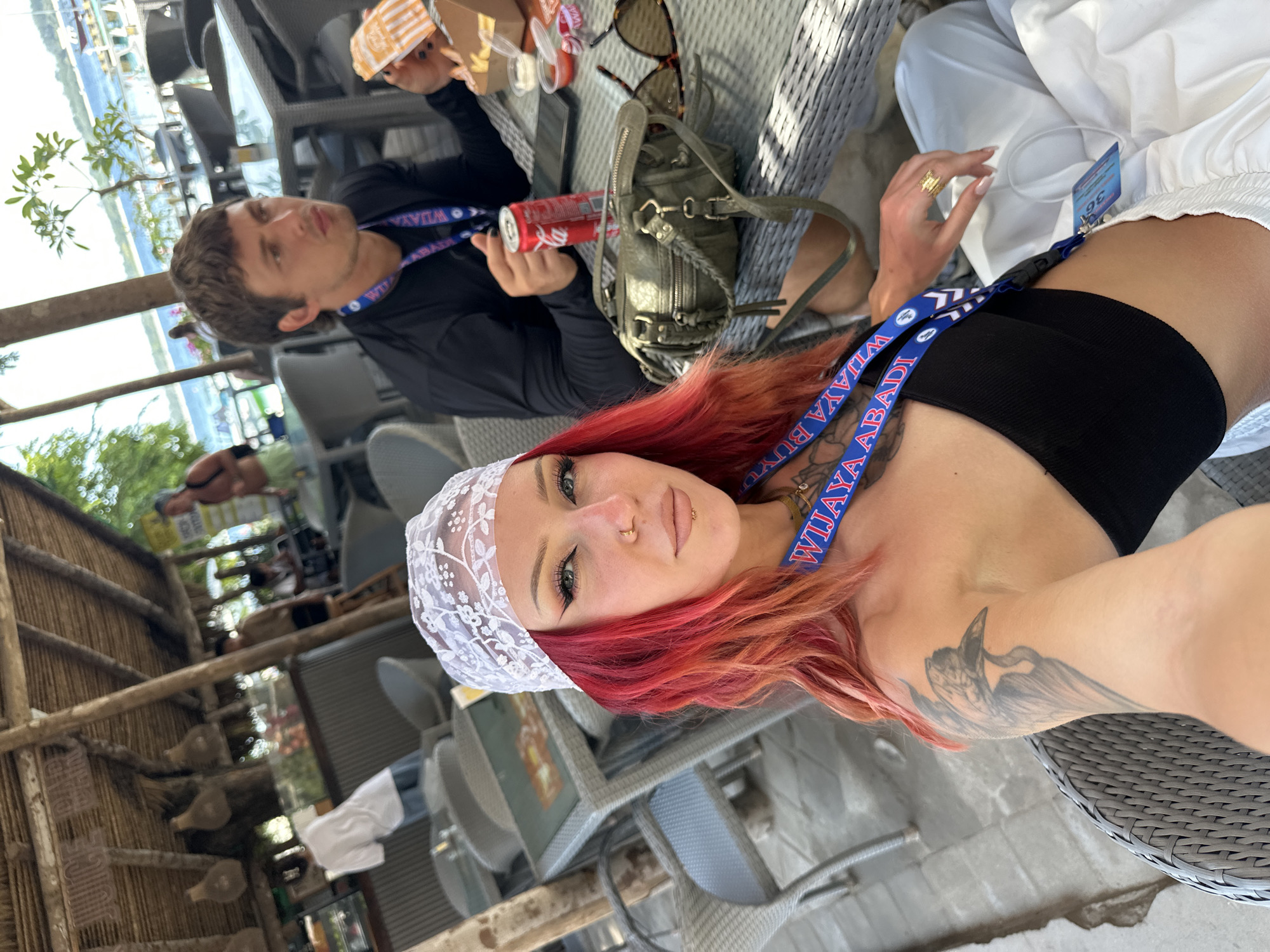 The scene depicts a selfie of a woman with red hair and a white lace headband at an outdoor dining area, likely near a beach. She has a tattoo on her arm and is wearing a lanyard with the words "WIJAYA ABADI" on it. Seated beside her is a man wearing a similar lanyard and a black shirt, holding a Coca-Cola can and a food container. A green handbag and sunglasses are on the table. In the background, there's a glimpse of a beach and a building with the words "JUICE BAR" visible. The setting appears to be a tourist spot in Unknown, Indonesia, judging by the scenery and the promotional lanyard.