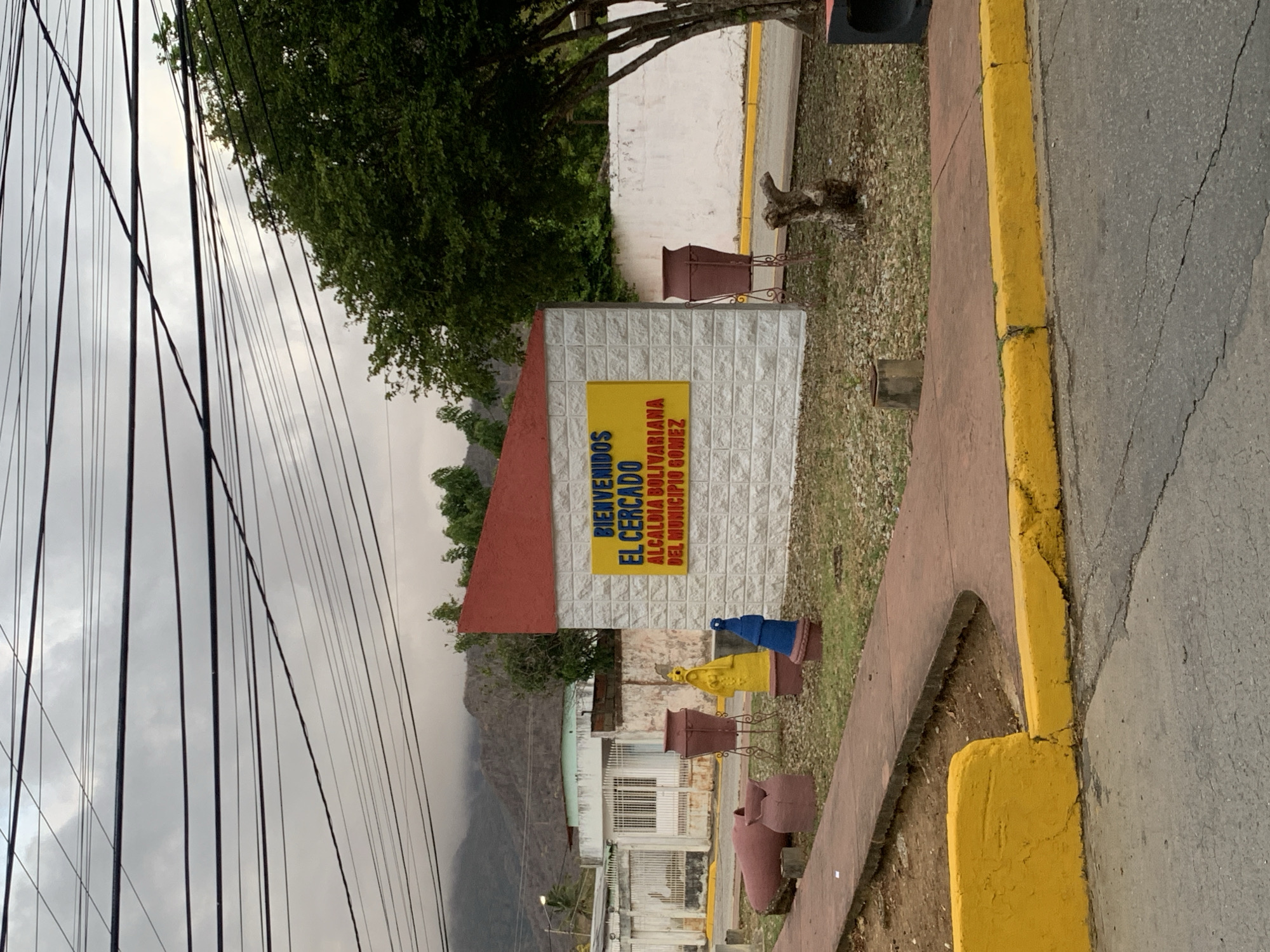 The image displays an outdoor scene, likely a roadside entrance to a locality, featuring a welcome sign. The sign, mounted on a white, textured block wall with a reddish-brown angled roof-like top, is prominently yellow with red uppercase text. It reads: "BIENVENIDOS EL CERCADO ALCALDIA BOLIVARIANA DEL MUNICIPIO GOMEZ."
In front of the sign structure and extending to the right, several decorative pots are visible on a grassy, dirt-covered area; some are reddish-brown, one is blue, and another is yellow. To the right, a bright yellow curb separates a reddish-brown concrete path from an asphalt road that shows minor cracks. Beyond the sign, hints of modest buildings or houses can be seen.
The background features lush green trees and a range of mountains or hills, suggesting a rural or semi-rural setting. The sky is heavily overcast with diffused light, indicating a cloudy day, possibly early morning or late afternoon. Numerous black utility wires crisscross high above against the grey sky. No people or specific activities are visible. The overall scene suggests a typical entrance to a town or village in a mountainous region of La Tagua, Venezuela.