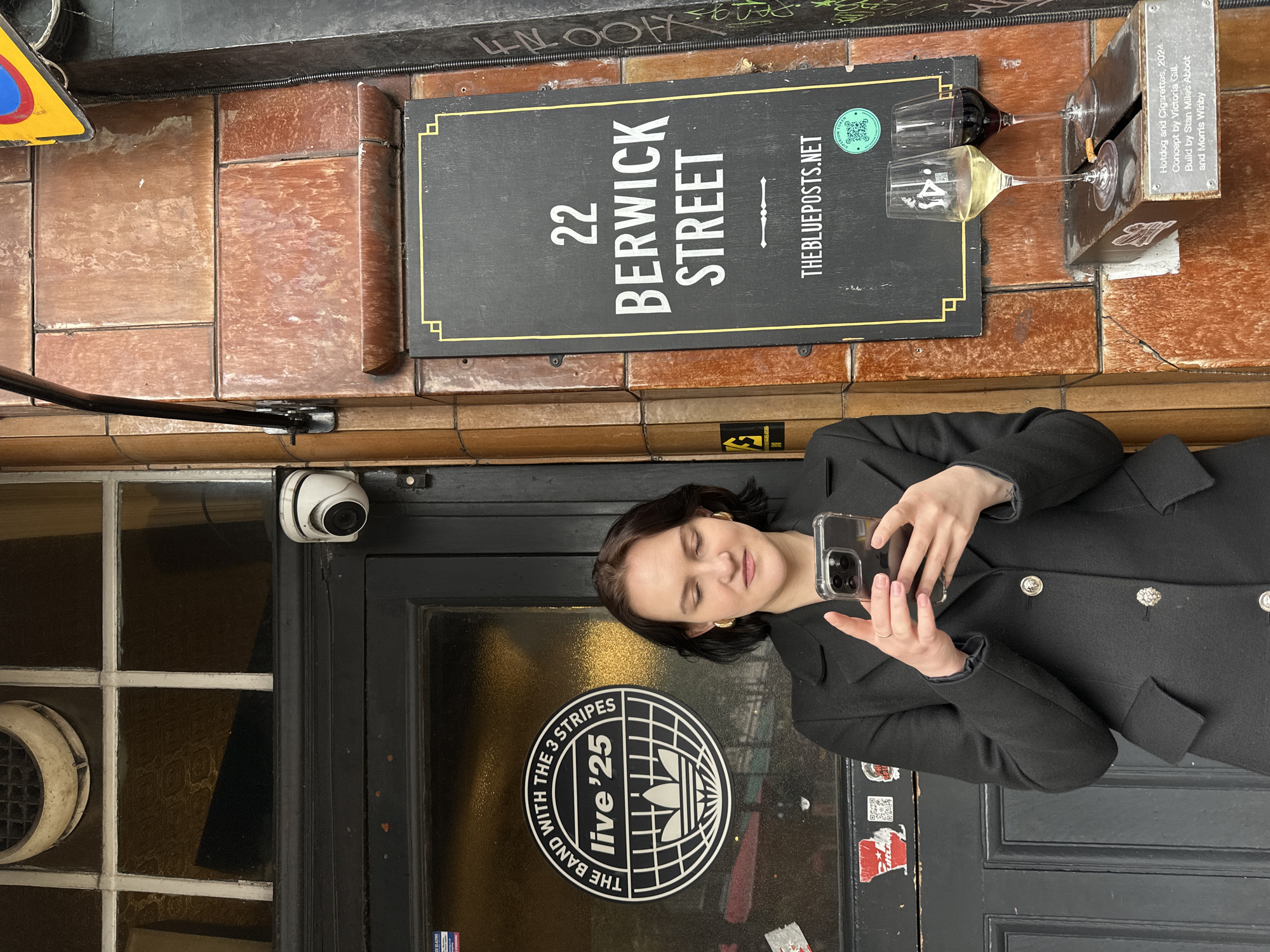 An urban exterior scene on Berwick Street in Soho, London, United Kingdom, features a young adult female and various architectural and artistic elements. The woman, with short dark hair and wearing a black double-breasted coat with metallic buttons and gold hoop earrings, stands centrally, looking down at a smartphone held in both hands. Her fingernails are painted light pink.
To her left is a dark wooden entrance door with recessed panels. Above the door, a multi-pane window features a circular vent at its top. A white dome security camera is mounted above the doorframe. On the glass of the door, a prominent circular black and white sticker displays "THE BAND WITH THE 3 STRIPES live '25" enclosing an Adidas globe logo. Several smaller stickers are visible below it, including a red and white one and a QR code.
To the woman's right, the building's facade is constructed from reddish-brown rectangular tiles, showing signs of weathering. A tall, vertical black sign with a yellow geometric border is affixed to this tiled wall. It reads "22 BERWICK STREET" in white capital letters, followed by a decorative arrow motif, and "THEBLUEPOSTS.NET" at the bottom. A circular green sticker with a QR code and the words "PREMIUM TOKEN" is placed near the bottom right of the sign.
Below the sign, a dark metal box or shelf protrudes from the wall. Two stemmed wine glasses are placed on its top surface: one containing white wine on the left, and one with red wine on the right. A single cigarette butt rests inside the white wine glass. A rectangular plaque on the front of the metal box is engraved with the text: "Hotdog and Cigarettes. 2024. Concept by Victoria Gill. Build by Stan Miles Abbot and Morris Winby." The far right edge of the image reveals a dark, textured wall with some indistinct graffiti. The lighting is even, suggesting an overcast day.