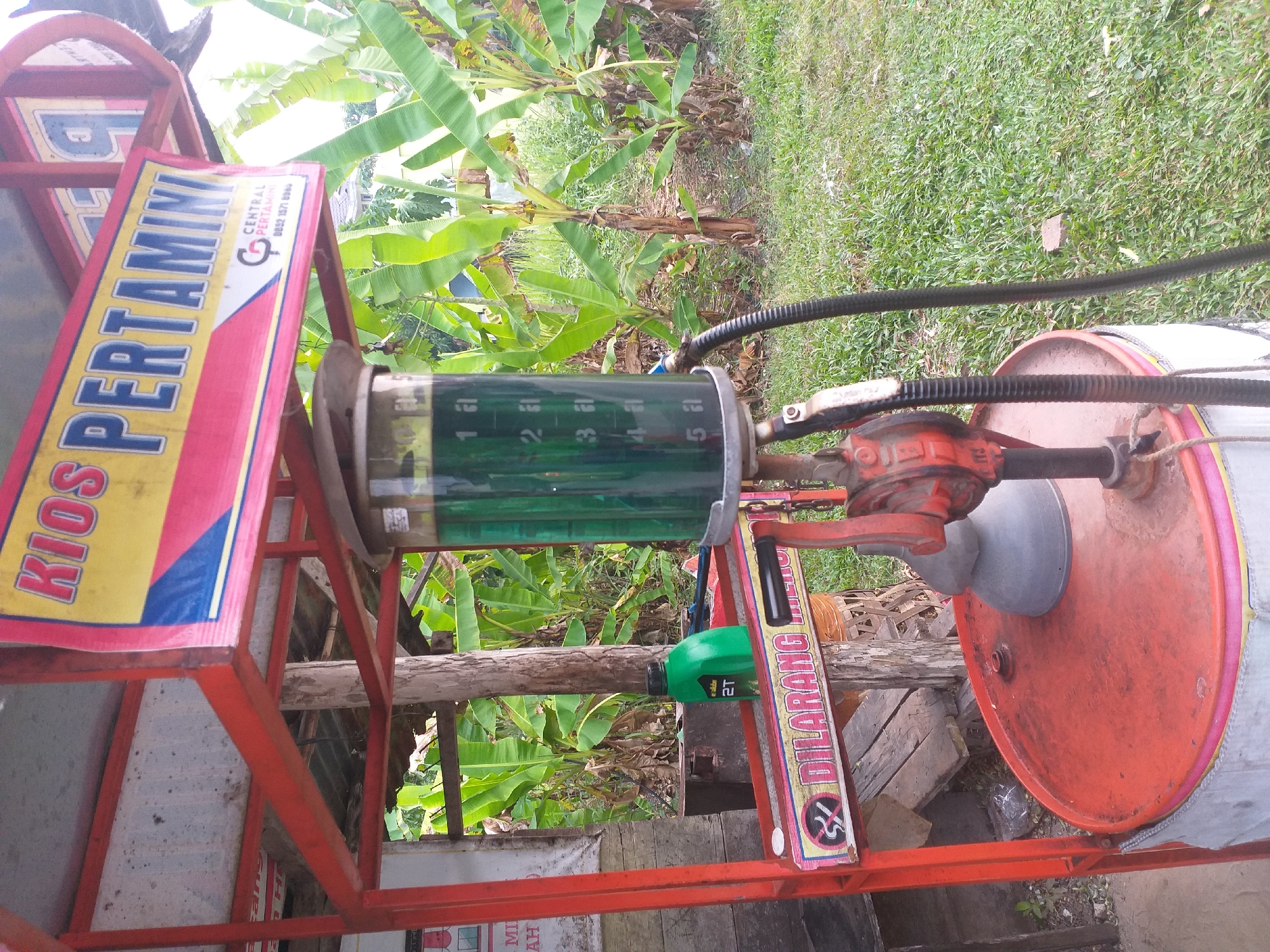 The image depicts a roadside fuel pump, commonly known as a "Pertamini," located in the city of Air Jamban, Indonesia. The pump is constructed with a red metal frame and features a transparent cylindrical tank filled with green fuel. A black hose extends from the pump mechanism to the ground. A red manual pump handle is attached to the side of the pump. Below the fuel tank, a sign reads "DILARANG MEROKOK," which translates to "NO SMOKING," accompanied by a crossed-out cigarette symbol. A green bottle with a black cap is visible on a shelf to the left of the pump, with the label "2T" clearly displayed. In the background, lush green banana trees and vegetation suggest a tropical environment. The ground is covered in grass. The overall scene suggests a functional, albeit rustic, fuel dispensing station in a rural or semi-rural Indonesian setting.