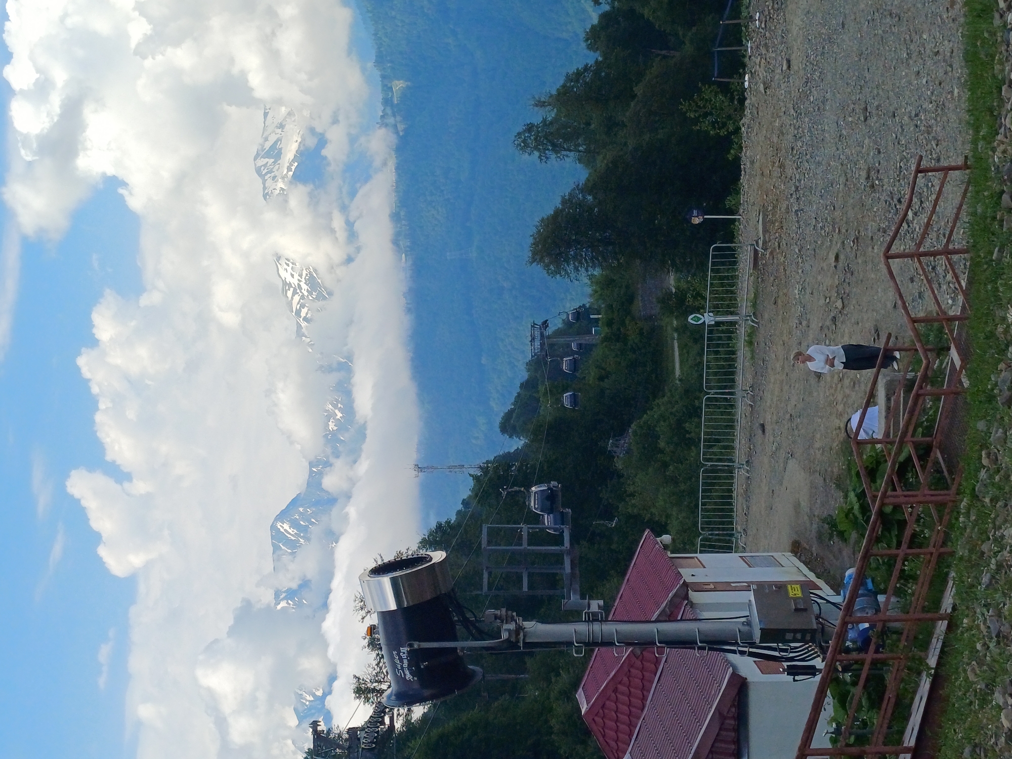 The image captures a mountain landscape with a ski lift in operation. In the foreground, a large, dark snow cannon sits atop a building with a red tiled roof. The cannon has silver accents and text that reads "Super" and "Defroster". To the right, a person in a white top and dark pants stands near a rusty metal railing overlooking a gravel path. In the mid-ground, the ski lift with several cabins can be seen ascending a forested mountain slope. Beyond the trees, snow-capped mountains rise into a sky filled with dramatic white clouds. The overall scene suggests a daytime setting, likely during warmer months given the lush green foliage on the lower slopes. The vastness of the mountains and the presence of the ski lift hint at a resort area in a mountainous region, consistent with the location context of Esto-Sadok, Russia.