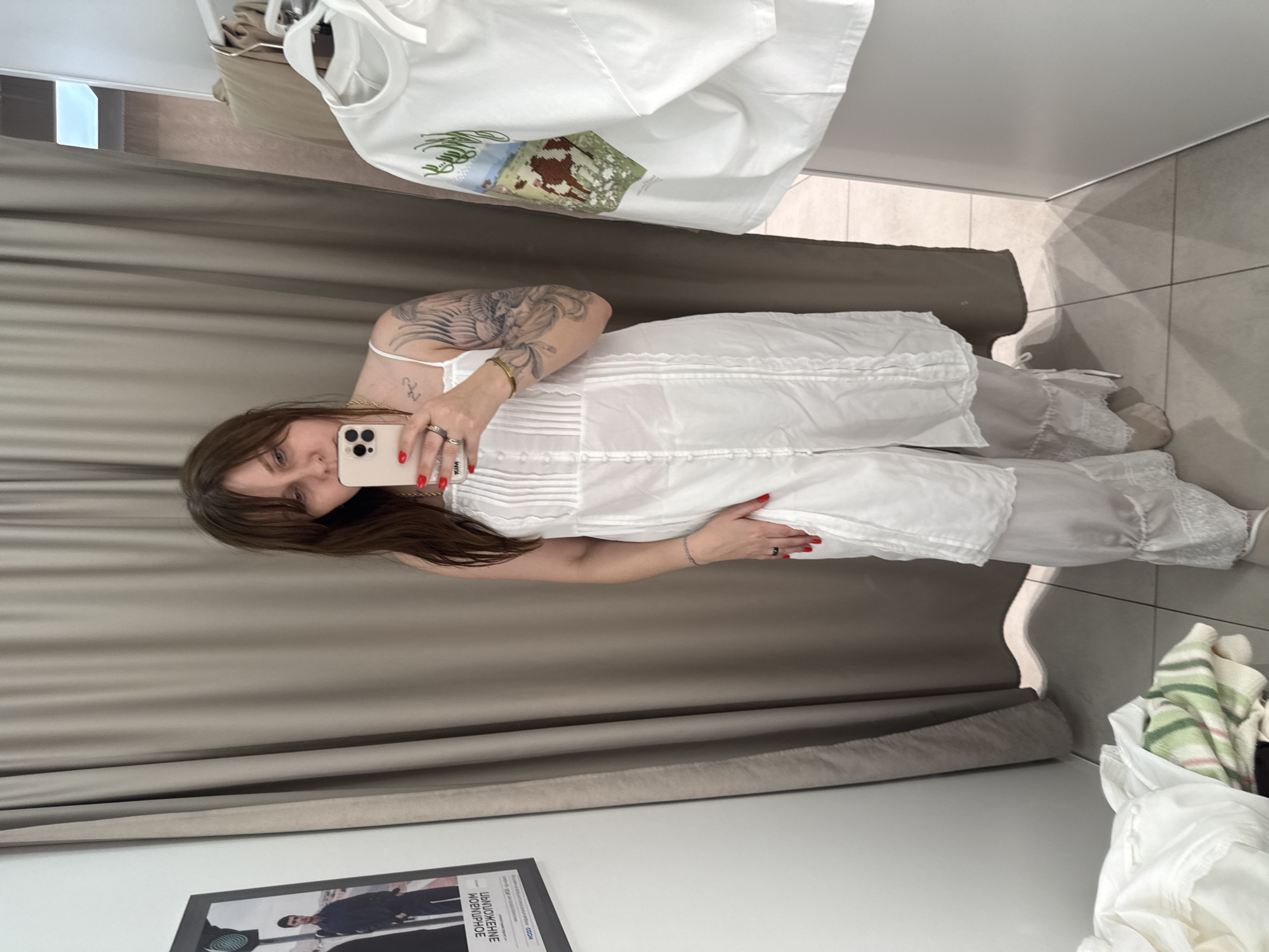 In a fitting room with beige curtains and tiled flooring, a woman stands in front of a mirror, taking a selfie with her phone. She has long, dark brown hair with bangs and is wearing a white slip dress over white pants with lace trim. She has tattoos on her arms and is wearing rings and a bracelet. To her right, a white t-shirt with an embroidered image of cows in a field hangs on a hanger. A framed poster of a man in a dark jacket and pants is visible on the wall to her left. A pile of clothes is on the floor in the lower left corner. This scene is located in a fitting room, which could be found in a retail store in Sochi, Russia.