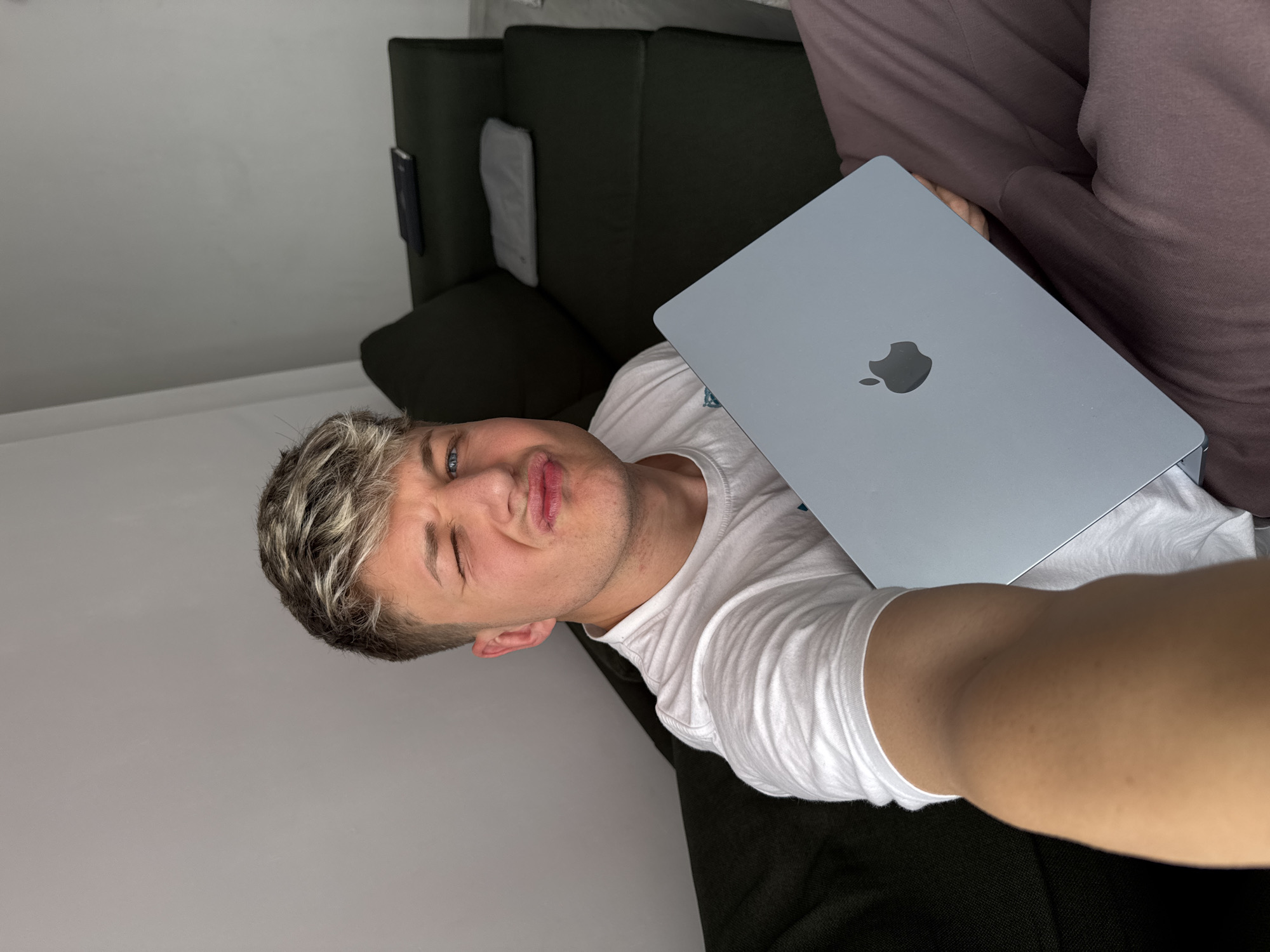 A selfie of a young man with short, bleached hair lying on a dark green couch while holding a closed, silver MacBook. The man is wearing a white T-shirt and is making a playful, squinting face at the camera. He appears to be indoors, with a light gray wall visible in the background. There is also a person wearing brown trousers to the top-right of the image. It is not possible to determine the location as being Plovdiv, Bulgaria from the image.