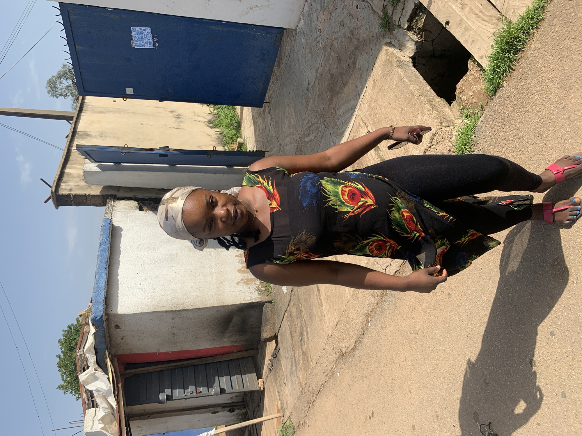 In Jos, Nigeria, a young woman with dark skin and braided hair covered by a patterned headscarf is captured mid-stride, facing the camera. She wears a black sleeveless dress adorned with colorful feather-like patterns, paired with black leggings and pink flip-flops. Her right hand holds a red mobile phone. The background reveals a narrow, unpaved street with a concrete drainage ditch on the right side. Adjacent structures include a small, white-washed building with a red-framed doorway and a corrugated metal roof covered with white sheeting, and a larger, cream-colored wall with two tall blue gates. The overall environment suggests a residential or commercial area with modest infrastructure.