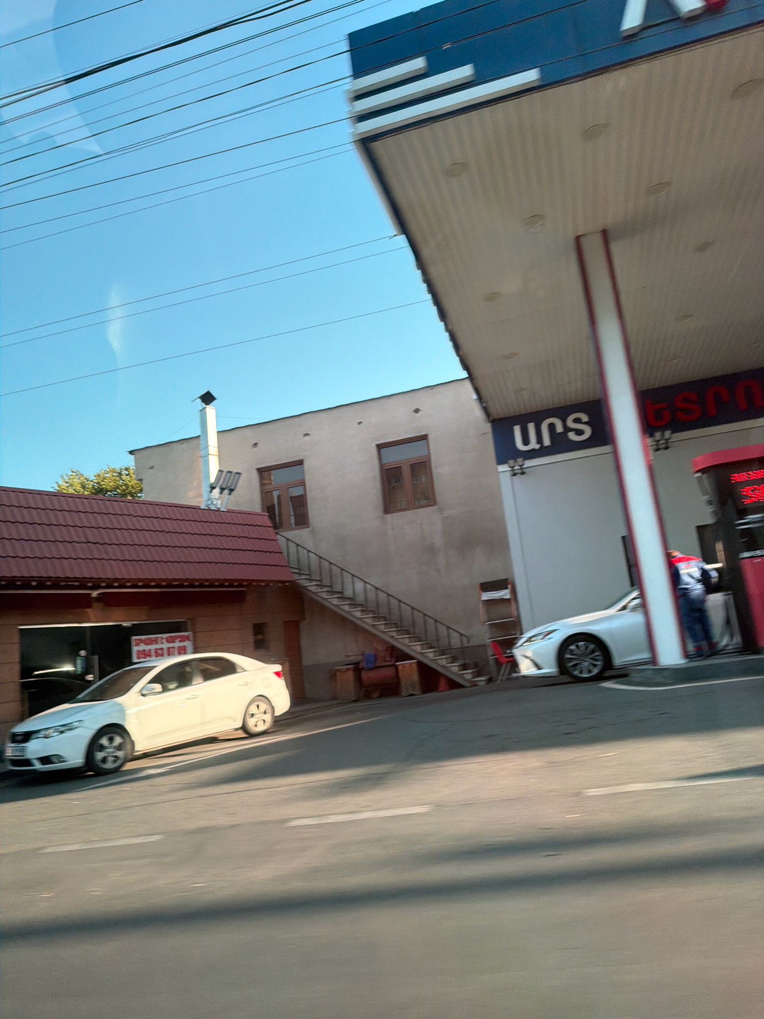 The image captures a street scene in Yerevan, Armenia, likely near a gas station. In the foreground, a light-colored sedan is parked on the right side of a road marked with white lines. A second white sedan is positioned in front of a building with a red-tiled roof. This building appears to house a business, with a sign displaying Armenian text and a phone number "094 63 17 09".
Behind the building with the red roof, a taller, light-colored structure with brown-framed windows and an external staircase is visible. Above this building, a metal chimney with attached lights extends upwards.
To the right, a gas station canopy extends over the street. The canopy is supported by a white pillar with red trim and features blue and white signage with Armenian lettering. A white Lexus sedan is parked under the canopy, near a red gas pump structure. A person wearing a blue and red uniform is standing near the gas pump, possibly attending to a customer or performing duties. The sky above is clear and blue, with several horizontal power lines stretching across it.