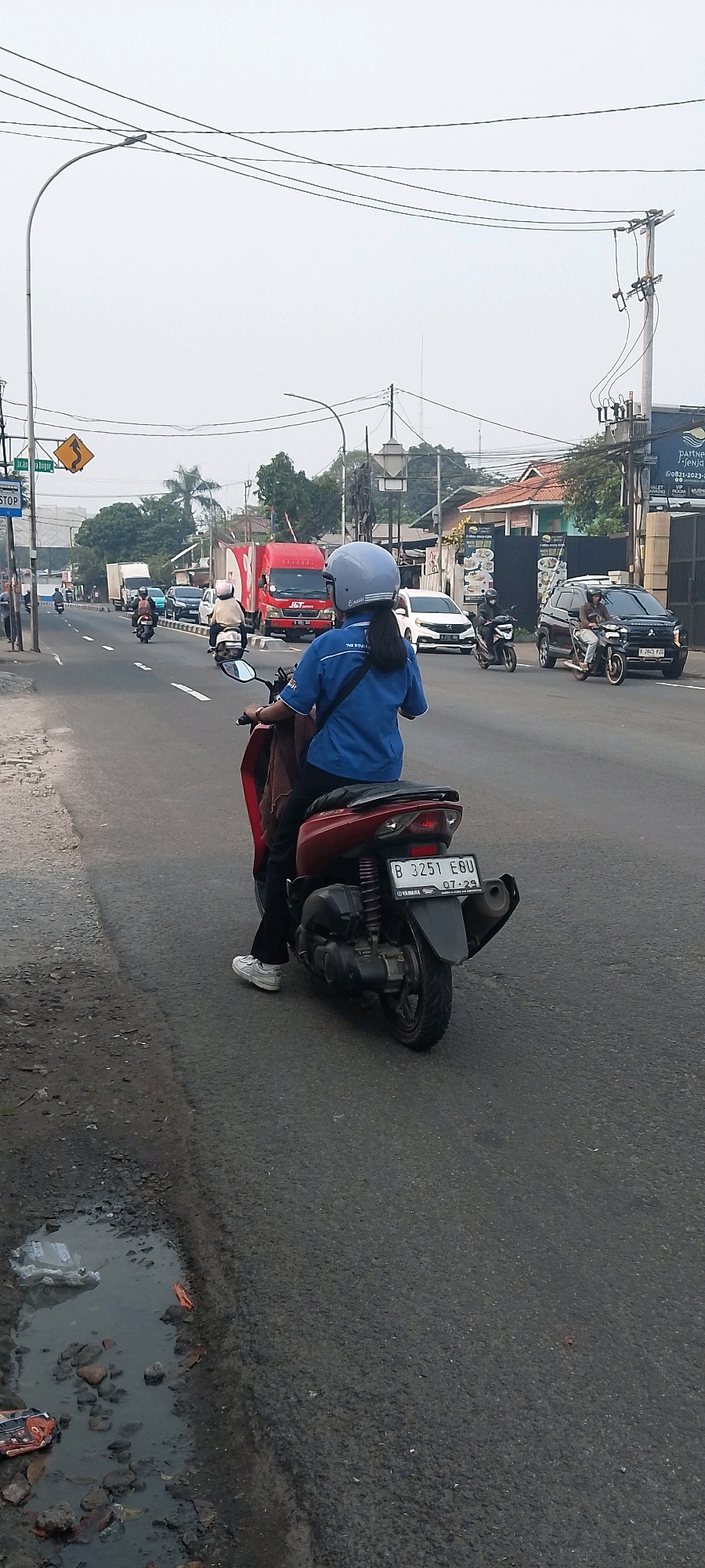 A woman wearing a blue shirt and a light blue helmet is riding a red scooter on a city street in Depok, Indonesia. She is in the foreground, facing away from the camera and looking towards the road ahead. The scooter has a license plate that reads "B 3251 EGU" and a sticker indicating "07-29". The road is paved and appears to be a busy street with several other vehicles, including cars and motorcycles, in the background. There are also traffic lights, street signs, and buildings lining the street. The sky is overcast, suggesting it might be a cloudy day. The side of the road on the left has a damaged pavement and a puddle of murky water with some debris.