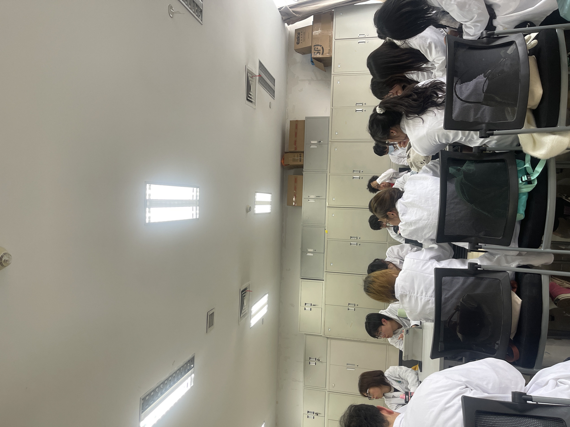 This image depicts an indoor meeting or classroom setting located in Yuzhong District, China. Approximately 15 to 20 individuals are visible, all wearing white lab coats and seated in black mesh-backed chairs at light-colored tables. Most individuals are oriented towards the front of the room. One person is observed with a tablet device on a table. The background wall features multiple rows of uniform light-grey metal storage cabinets with double doors and handles. Several cardboard boxes are stacked on the uppermost cabinets, with one box prominently displaying the "SF" (SF Express) logo, indicating a Chinese operational context. The ceiling is white, equipped with multiple rectangular fluorescent light fixtures, square air vents, and a circular smoke detector. The upper wall section above the cabinets exhibits some surface irregularities and discoloration. The room is brightly illuminated by artificial lighting.