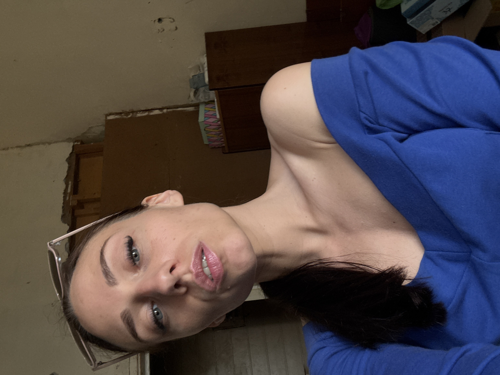 A woman with blue eyes and dark hair pulled back into a ponytail takes a selfie. She is wearing a royal blue off-the-shoulder top. Sunglasses are perched on her head. The background appears to be an indoor, somewhat neglected space with wooden furniture and pale, textured walls. The lighting suggests it is daytime, with light coming from the left. There are no discernible landmarks or features that would indicate a specific location within Saint Petersburg, Russia.