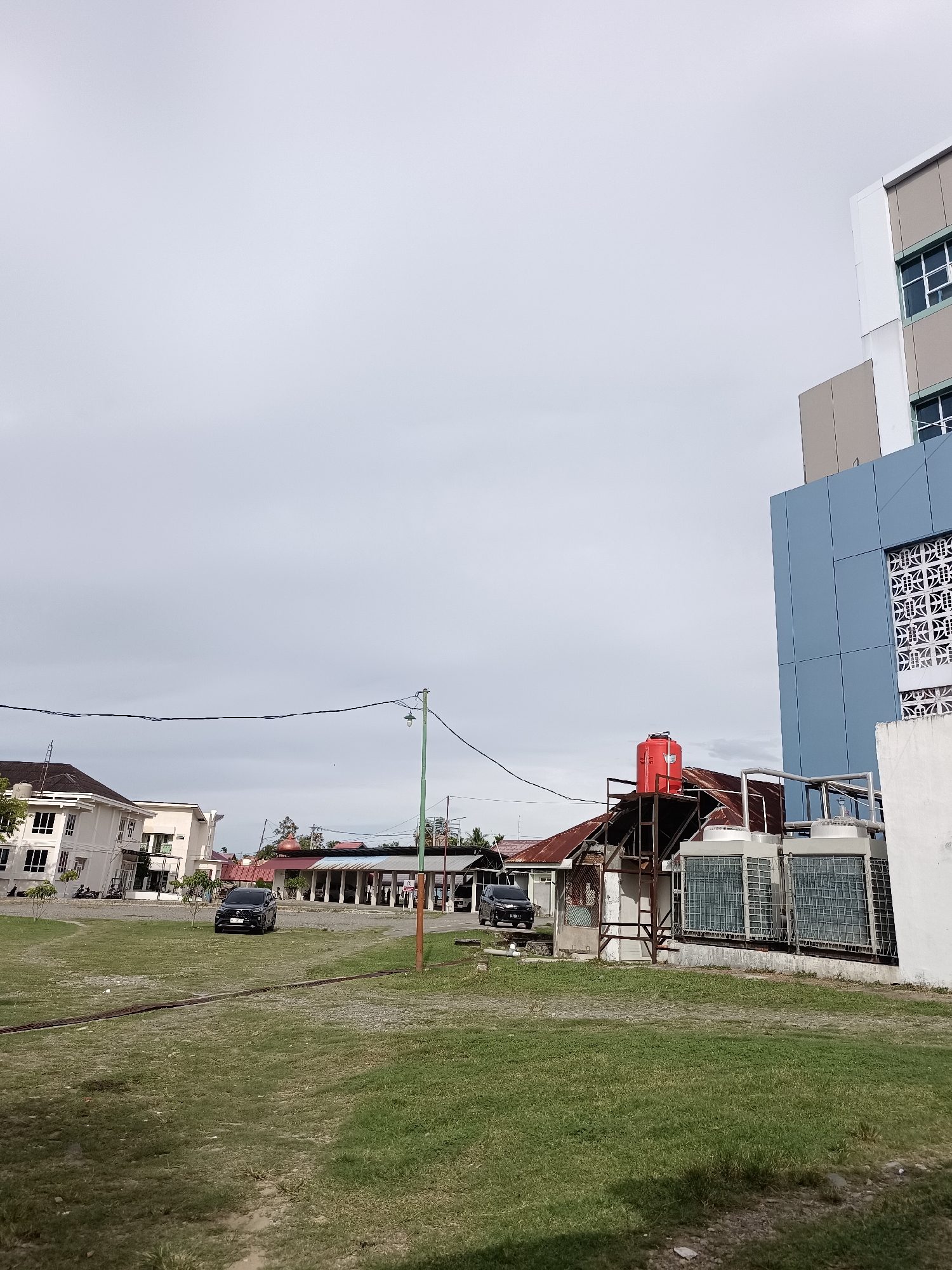 This is an eye-level, outdoor, medium shot of a building complex under a cloudy sky. The scene depicts an urban or semi-urban environment with a focus on architectural elements and infrastructure.
In the foreground, there is a grassy field with some gravel patches. Two black cars are parked in the mid-ground. Beyond the cars, there is a row of buildings with covered walkways and a few smaller structures, one with a red water tank on top of a metal frame. To the right, a modern, multi-story building with blue and beige facade panels is prominent. Large air conditioning units are visible on a platform attached to this building. A thin power line stretches across the upper portion of the frame, supported by a green pole.
The weather appears overcast, suggesting it might be early morning, late afternoon, or a generally cloudy day. There are no people visible in the frame, and the overall mood is quiet and still. The location is identified as Mesjid Reuntoh, Indonesia. No discernible text is visible in the image.