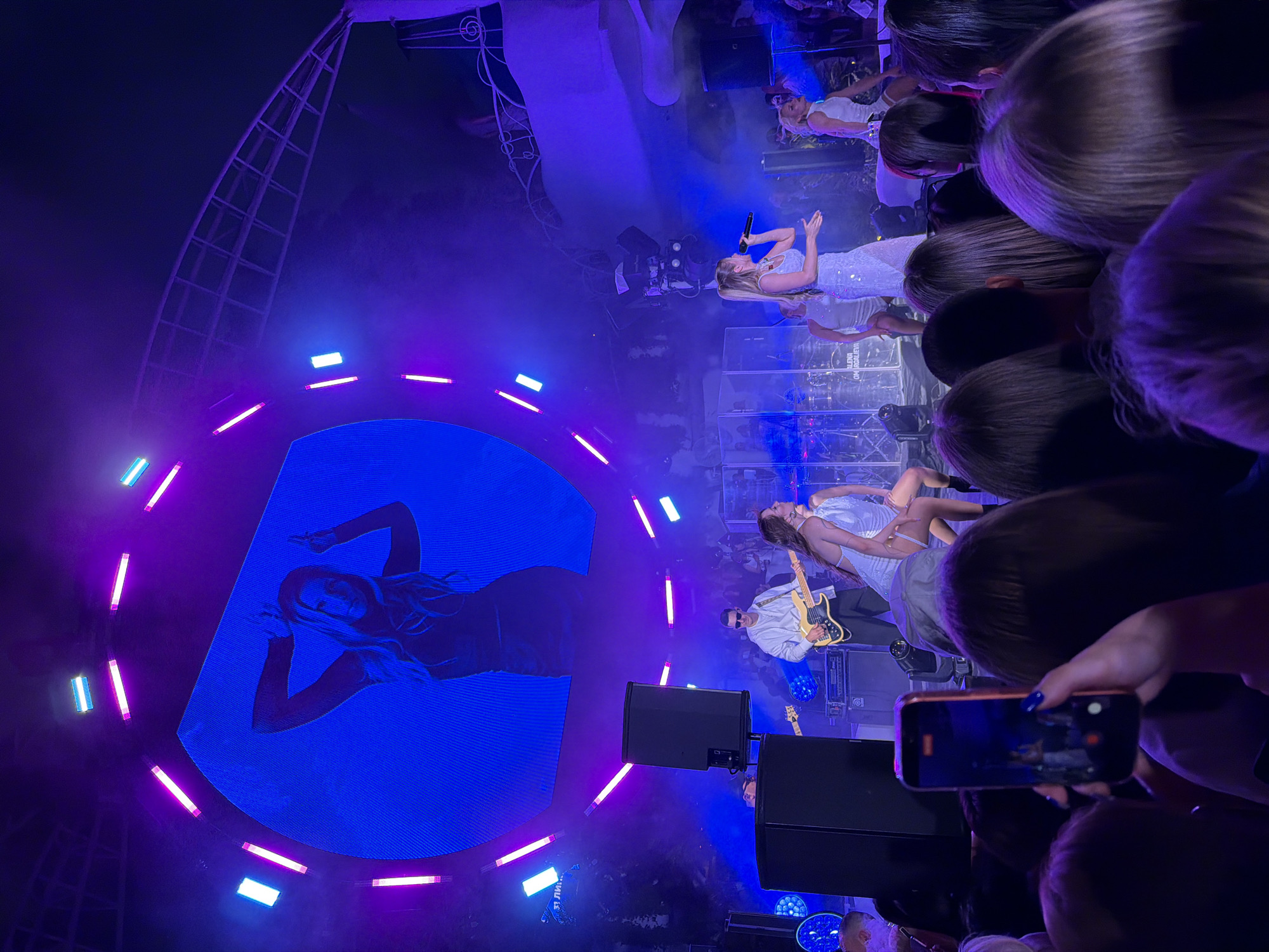The image depicts a concert performance at the "Ibiza Beach Club" in Odesa, Ukraine. In the foreground, the backs of several audience members' heads are visible, with one person in the center holding up a smartphone to record the event. The stage is illuminated with vibrant blue and purple lights, creating a dynamic atmosphere. A large circular LED screen dominates the background, displaying a silhouetted image of a woman with her hands raised. On stage, a female singer in a shimmering dress is holding a microphone and singing, with another female dancer to her right. A male musician is playing a bass guitar, and a drum kit is visible behind the performers. The overall scene is indicative of a lively outdoor concert at a popular beach club venue.