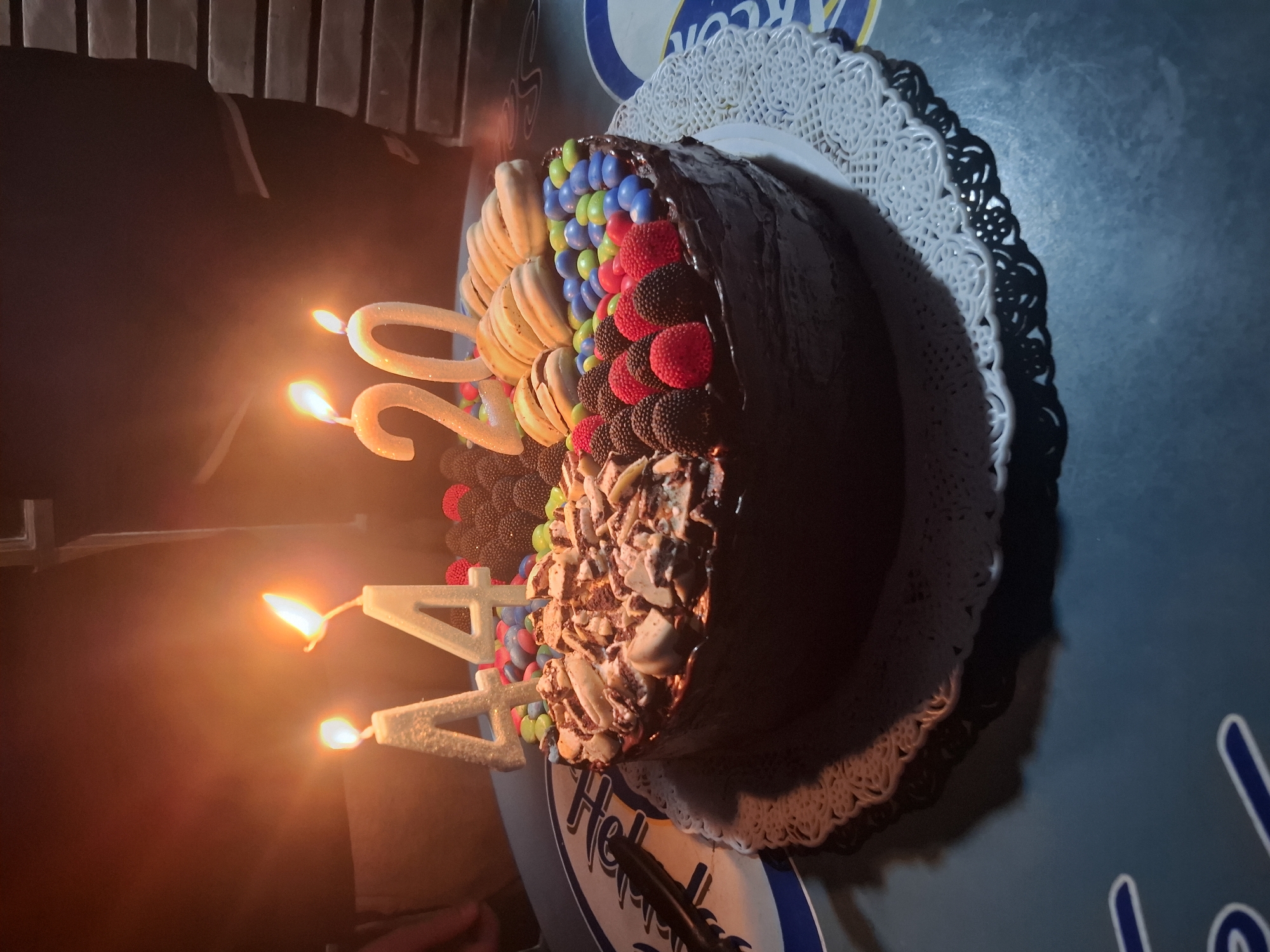 A birthday cake, decorated with candies, macaroons, and chocolate shavings, rests on a decorative plate with a scalloped edge. Two candles shaped as the number "4" and two candles shaped as the number "20" are lit, indicating a 44th birthday. The cake is placed on a surface with branding in the vicinity of Argentina, Buenos Aires. The scene is indoors and dimly lit.
