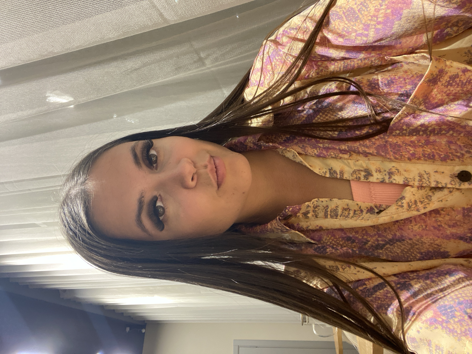 This media file is a portrait of a young woman with long, dark hair. She is wearing a colorful, patterned blouse with shades of yellow, pink, and purple. Beneath the blouse, a hint of a pink ribbed top is visible. Her makeup includes dramatic false eyelashes. The image is taken in an indoor setting, with horizontal blinds visible in the background, suggesting a window. The lighting appears to be artificial or from a window, creating a soft glow. The woman's gaze is directed slightly away from the camera, and her expression is neutral. The orientation of the image is portrait. No text is visible.