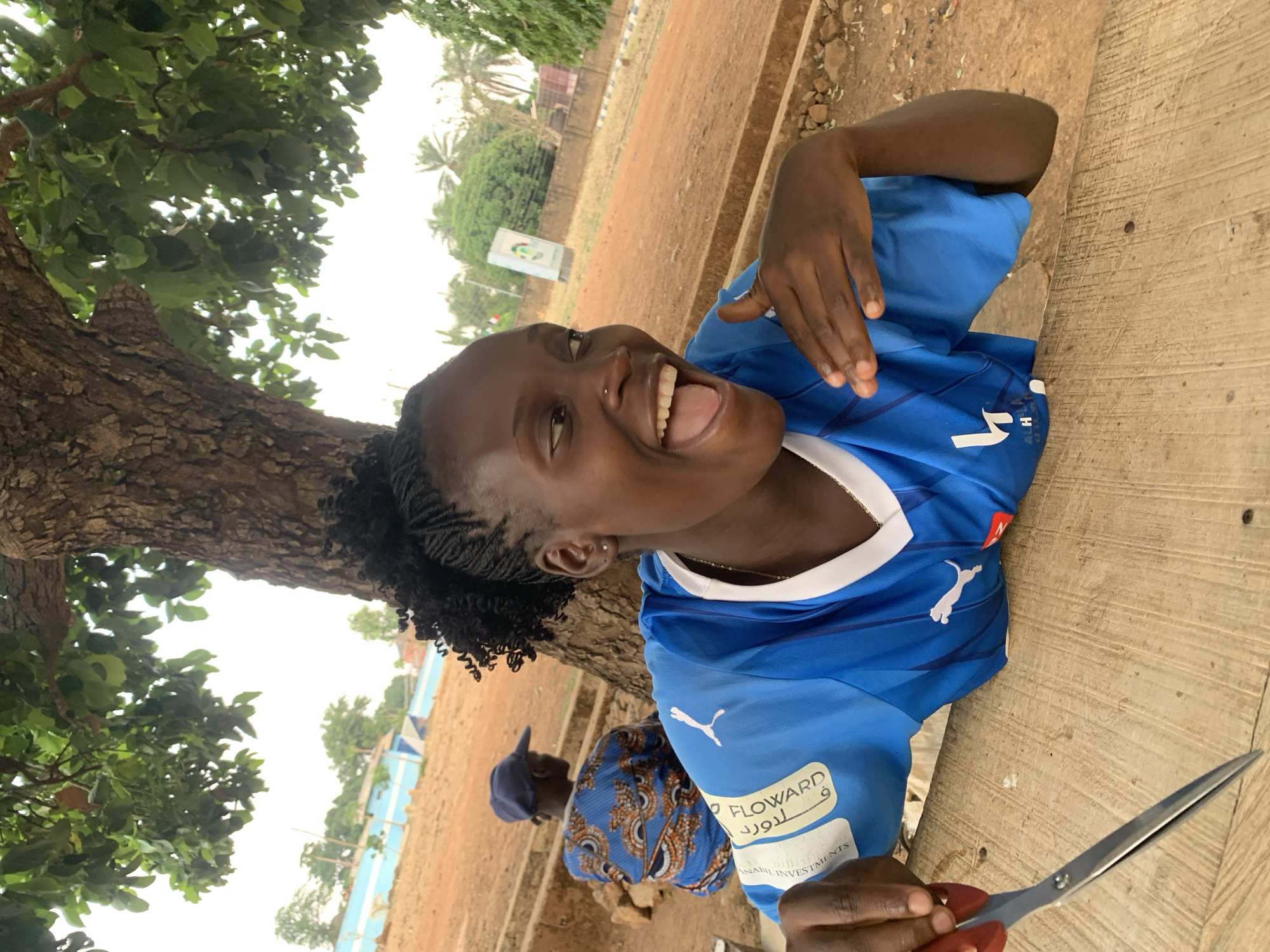 A young Black woman is photographed outdoors next to a tree. She is wearing a blue Puma soccer jersey with white trim and the logo for "Floward 9." Her hair is in braids, and she is smiling broadly, looking slightly upward to her left. She holds a pair of scissors in her right hand on a wooden surface. In the background, there is a glimpse of another person wearing a blue hat and patterned shirt. The surroundings suggest a park-like setting with dirt ground, trees, and foliage. Based on the prompt, the location is Maigizo, Nigeria.