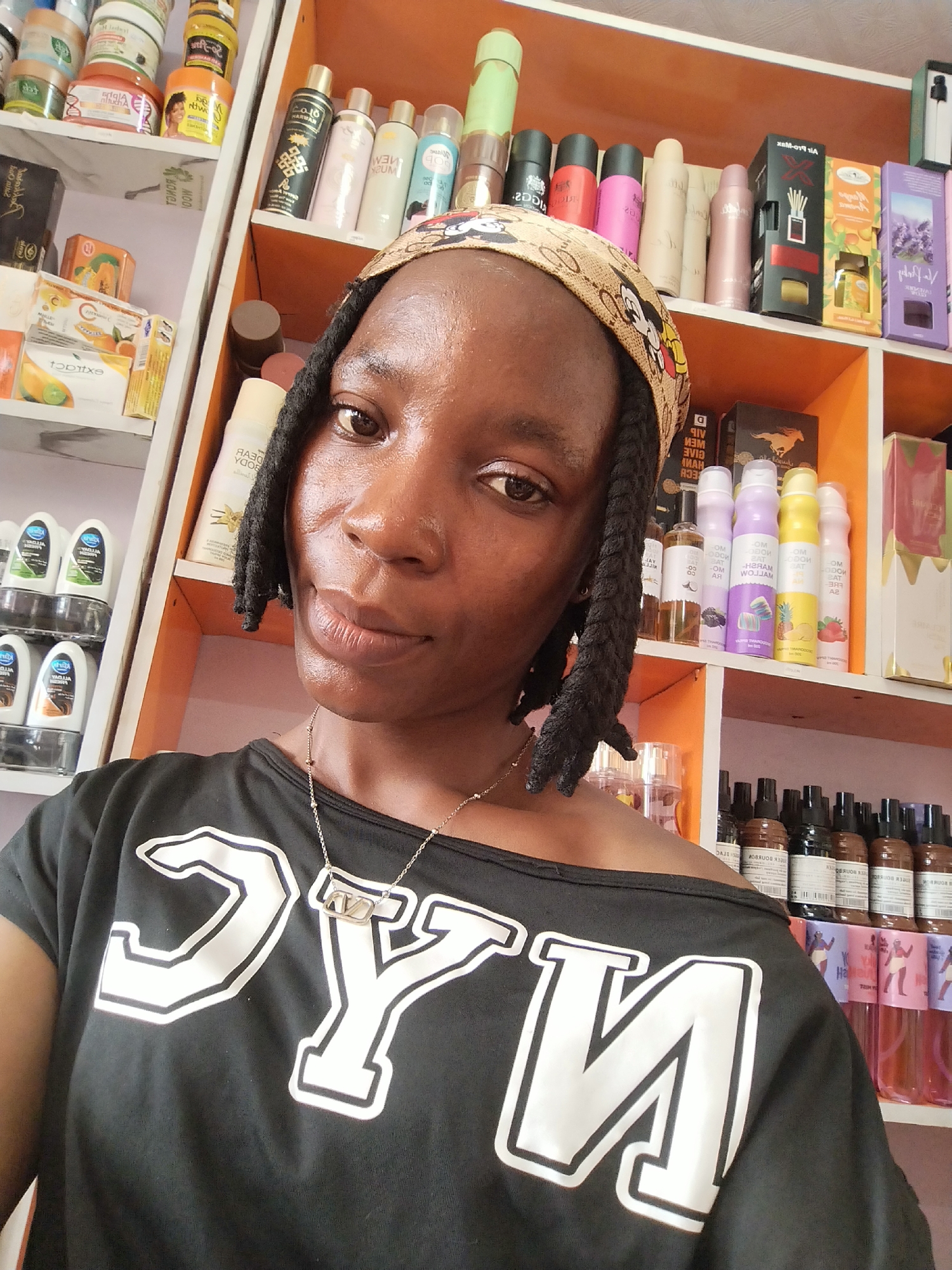 A woman of African descent, wearing a black off-the-shoulder shirt with a large white "NYC" graphic and a Mickey Mouse-themed bandana, takes a selfie in a retail store in Ugbowo, Nigeria. She has dark braided hair and is wearing a delicate silver necklace. The store shelves behind her are filled with an assortment of beauty products, including lotions, perfumes, and hair products. The shelves are painted orange and white, and the products are arranged in neat rows. The lighting is bright, suggesting an indoor environment.