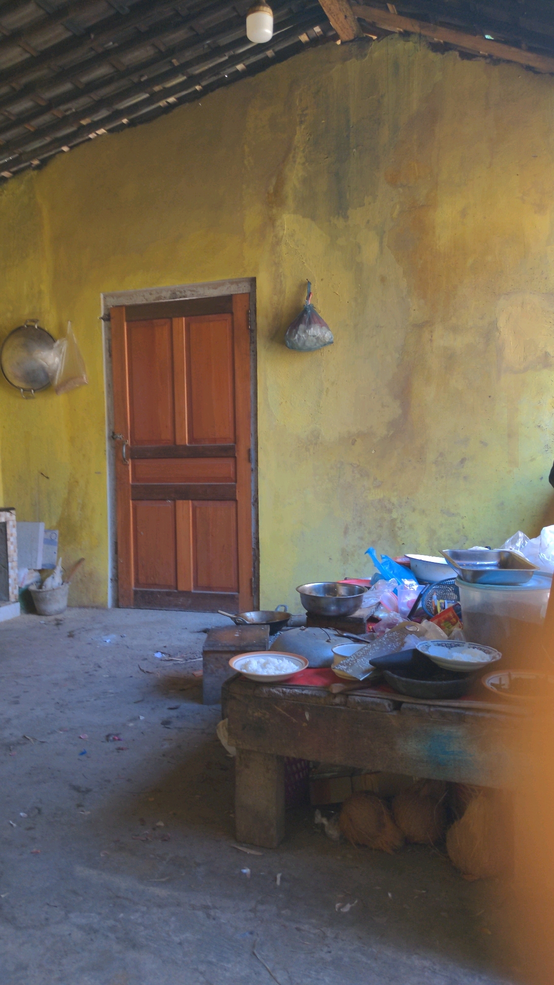The image depicts an outdoor cooking area in Tembarak, Indonesia. The scene is characterized by a rough-hewn wooden table laden with cooking implements, including bowls, plates, and what appear to be cooking vessels. The table is positioned in front of a weathered, yellow-painted wall. A wooden door is visible to the left of the wall. The ground is composed of concrete and appears somewhat dusty and littered. A roof structure, possibly thatched or tiled, is visible overhead, supported by wooden beams, and a simple light fixture hangs from the ceiling. Several coconuts are stored beneath the wooden table. The overall impression is one of a functional, perhaps rudimentary, kitchen environment.