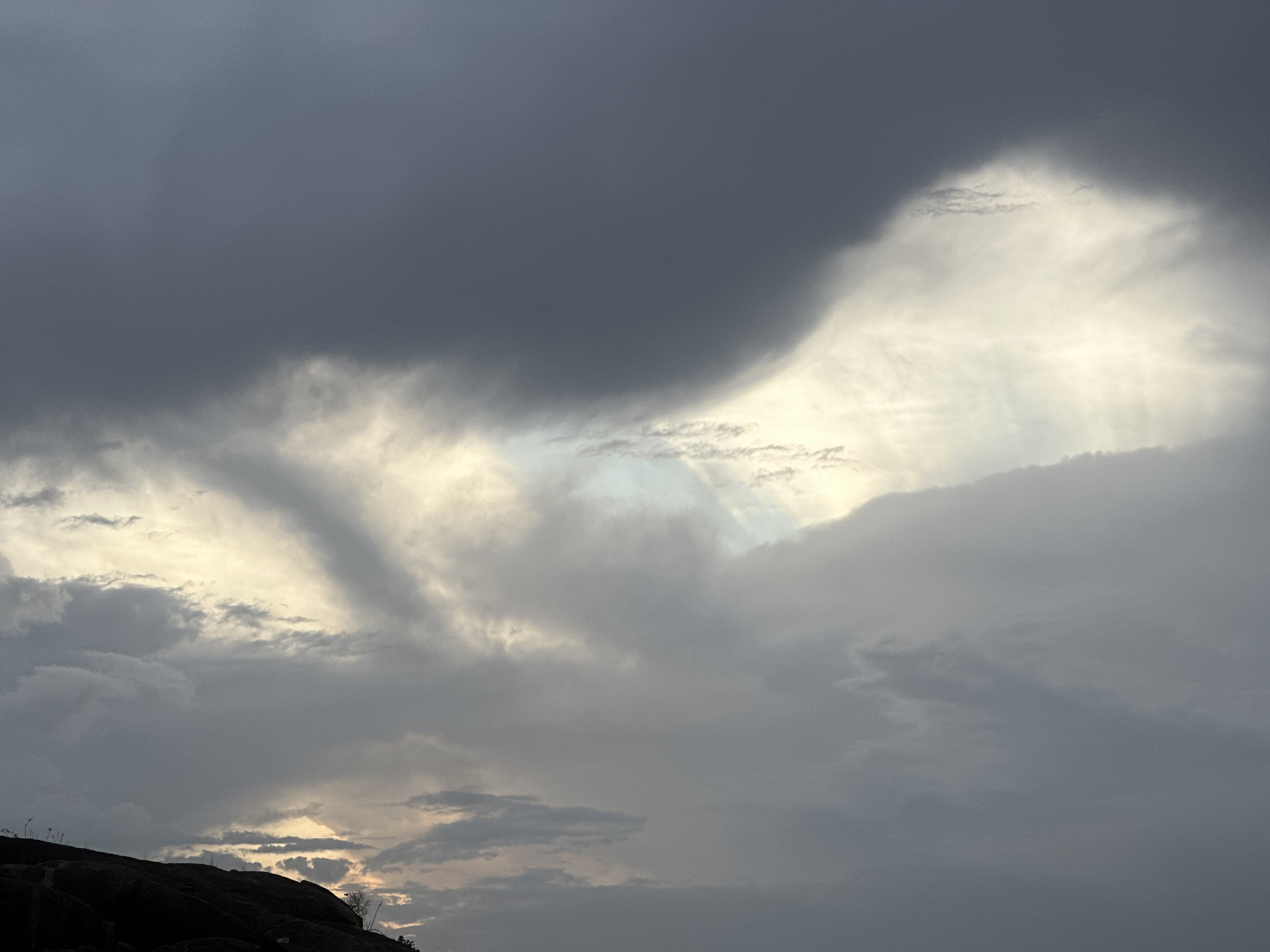 The image depicts a cloudy sky over a landscape in Jos, Nigeria. Dominating the view are heavy, dark gray clouds that fill the upper portion of the image. These clouds partially obscure the sky, with lighter, brighter patches visible where the sun manages to break through. At the bottom left corner, a dark, silhouetted mound or hill is present, likely a rocky outcrop typical of the Jos landscape. Sparse vegetation can be seen on this elevation. The overall mood of the scene is overcast, suggesting a potentially stormy weather condition.