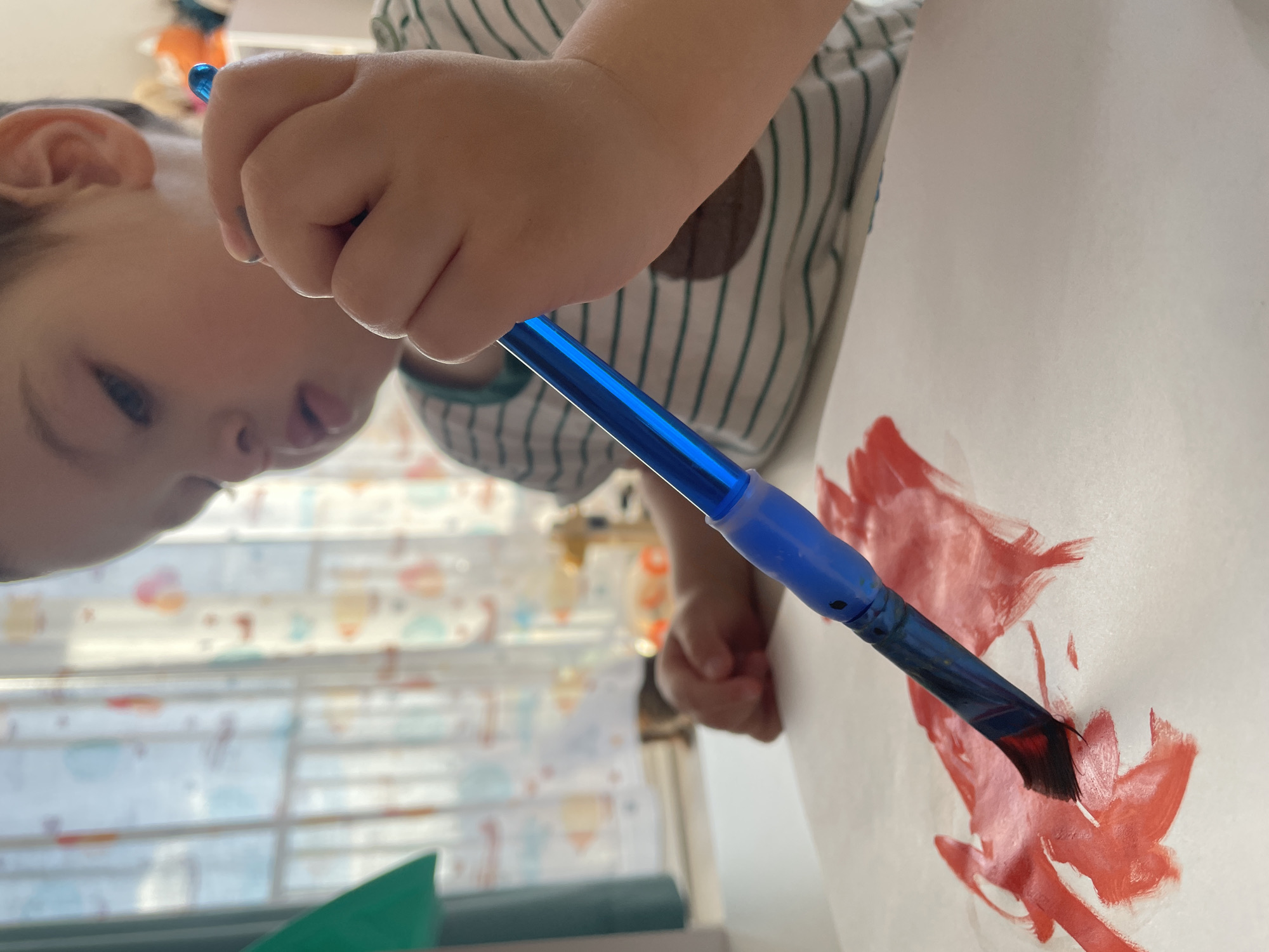 A young child is painting with a blue-handled brush on a white piece of paper. The child is wearing a vertically striped shirt with a round patch on the chest. Red paint is visible on the paper. The child's face is partially visible, showing their ear, eye, and mouth. A window with a patterned curtain is in the background. There is no information in the image to suggest the location is Izhevsk, Russia.