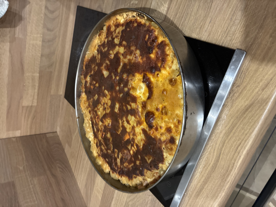 A circular, silvery metal baking pan holds a cooked dish, positioned on a black induction or ceramic stovetop. The dish's surface exhibits a varied texture, with areas of deeply browned, almost charred crust interspersed with lighter golden-yellow, likely creamy, sections. Small, light-colored diced ingredients are visible beneath the surface. The stovetop is integrated into a light-colored, wood-patterned laminate countertop. A matching wood-patterned backsplash forms a corner in the background. No individuals, actions, or direct interactions are depicted in the scene. The location is the city of Sofia, Bulgaria.