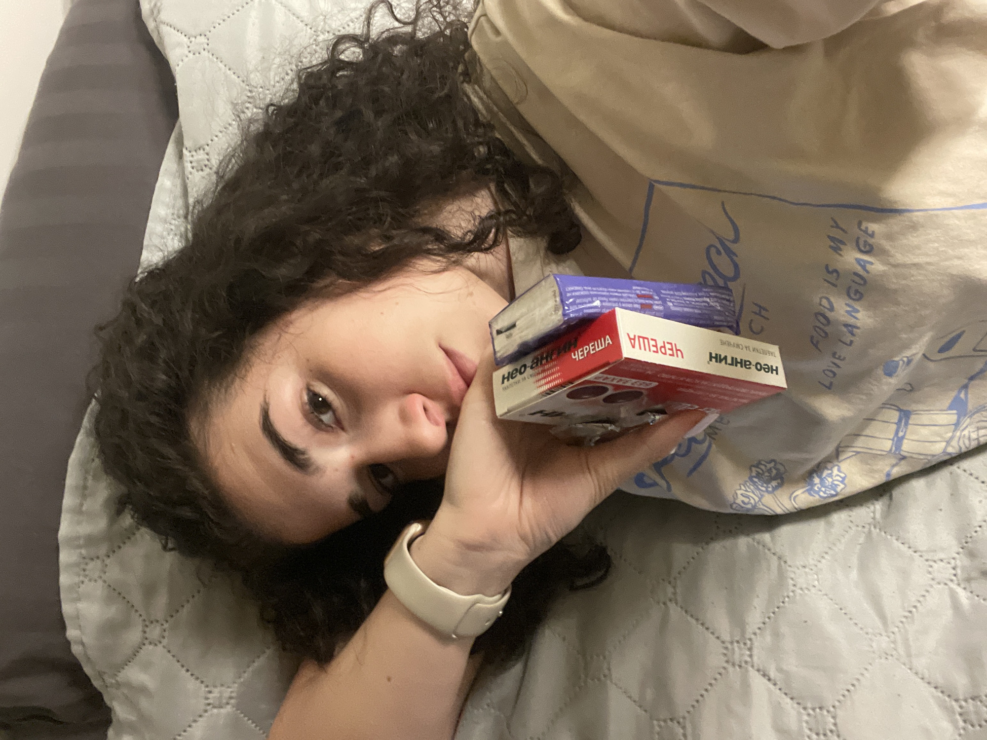 A woman is lying on a quilted gray bed cover and pillow, holding two boxes of "Neo-Angin" lozenges in her hand. One box is red with the flavor "Cherry" and the other is blue. The words "Neo-Angin" and "Cherry" are written in Cyrillic. She is wearing a beige t-shirt printed with "FOOD IS MY LOVE LANGUAGE." The exact location within the city of Sofia, Bulgaria is not discernible from the image.