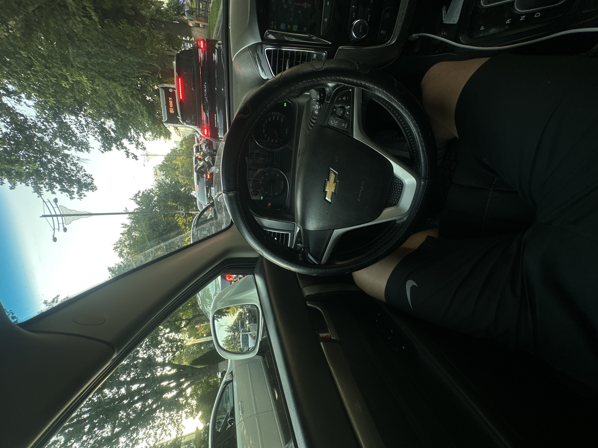 The image shows the interior of a Chevrolet car, with a person's legs visible in the driver's seat. The car is stopped in traffic on a street lined with trees. A black SUV is in front, with its brake lights illuminated. The dashboard displays a speed of approximately 0 km/h. The scene is set in Tashkent, Uzbekistan, as indicated by the license plate of the vehicle in front which reads "610HC". The sky is clear and bright, suggesting it is daytime.