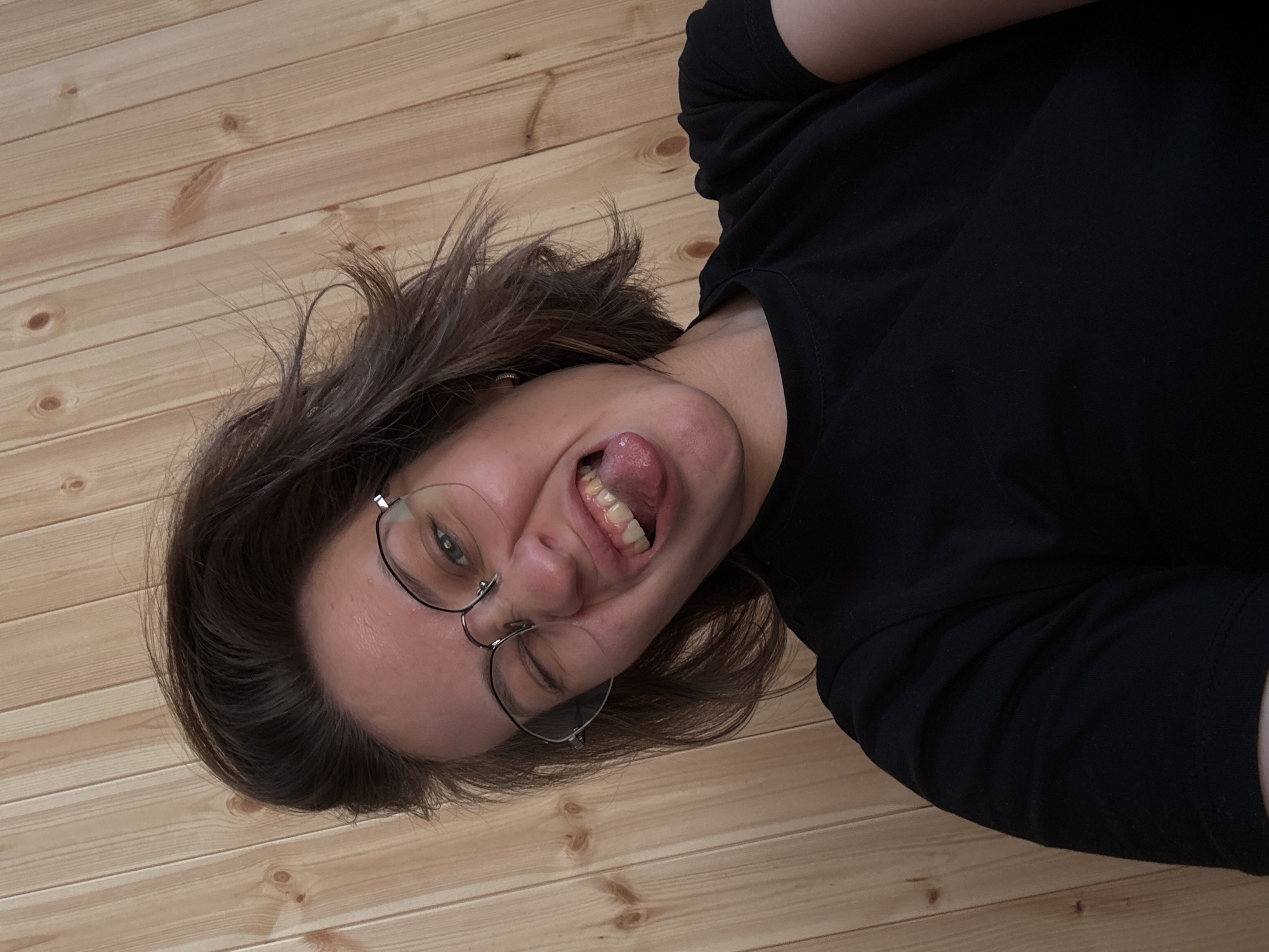 The image depicts an upward-angled selfie of an individual, likely in the city of Lenino, Russia. The subject is a person with short, dark brown, tousled hair, wearing clear-rimmed glasses with a metallic frame and a plain black crew-neck t-shirt. The individual is making a playful facial expression, characterized by a wink of the right eye, a protruding tongue, and a contorted mouth. A small, metallic hoop earring is visible in the subject's left earlobe. The teeth are visible, displaying a natural appearance with some minor yellow discoloration. The forehead shows a few small blemishes. The background consists entirely of vertically oriented, light-colored wooden planks, exhibiting natural grain patterns and knots, consistent with an interior wall paneling. The lighting is even and diffused.