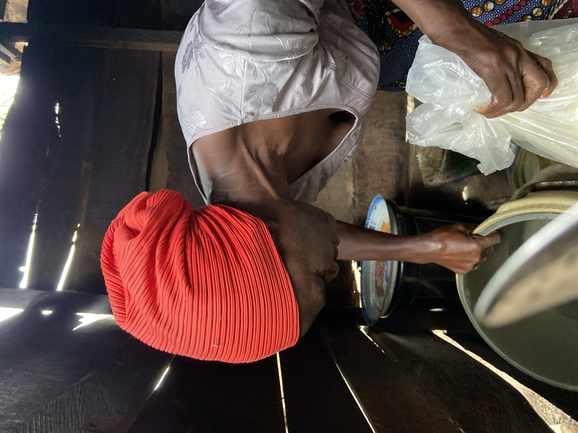 In Mararaba, Nigeria, a woman is captured indoors, standing near a wooden structure with light seeping through the slats. She wears a bright orange headwrap, a pale, floral-patterned covering around her head, and part of a patterned dress is visible. Her dark skin is visible. She holds a clear plastic bag in one hand and appears to be interacting with a metal basin or container with her other hand. There is a blue and white plate placed near the basin.