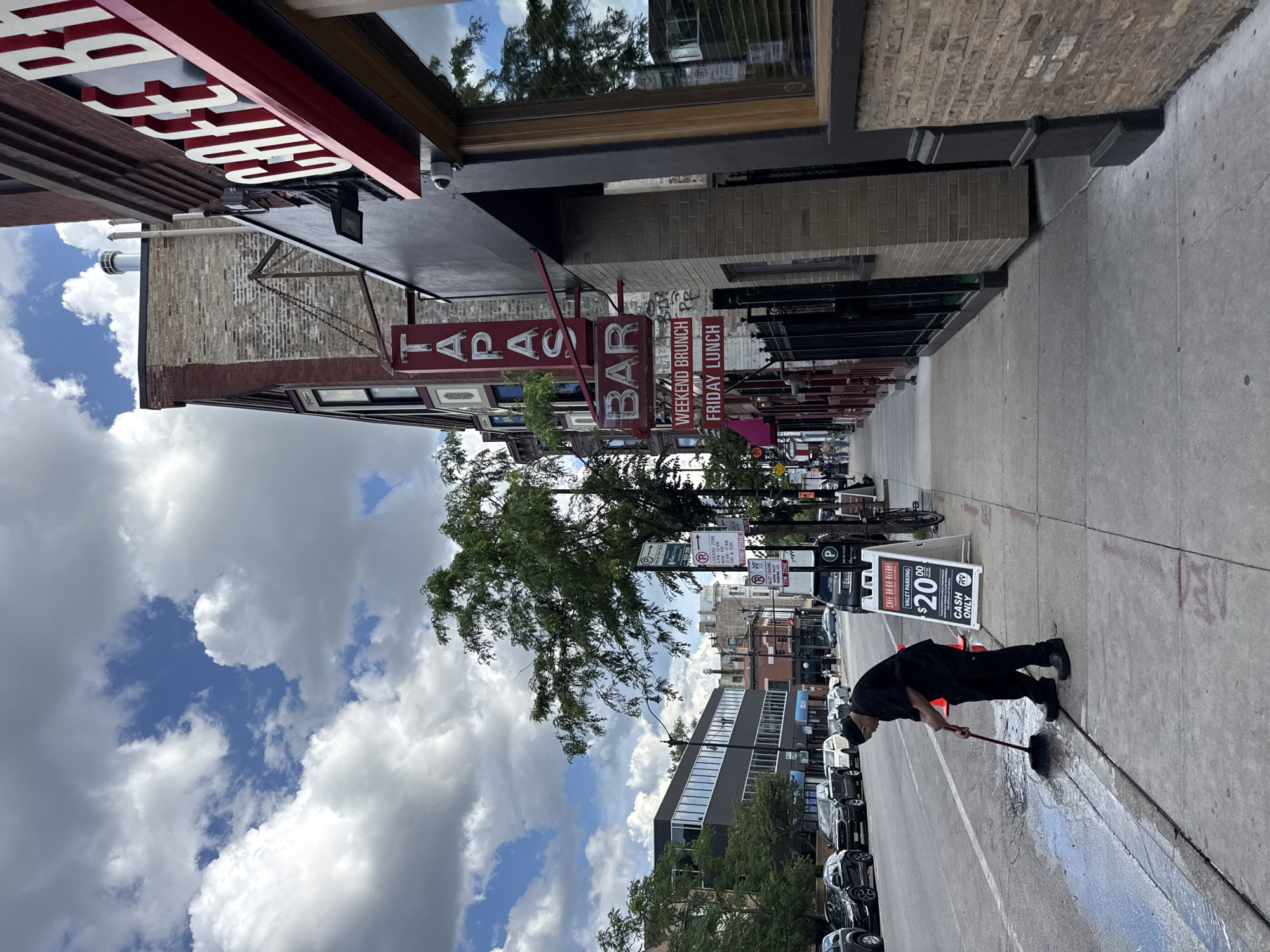 A man in all black clothing, including a beanie, is sweeping water on a sidewalk with a broom. He is near a sign that advertises "CAFE BA-BA-REEBA!" for "VALET PARKING $20.00 CASH ONLY". The sign also mentions "WEEKEND BRUNCH" and "FRIDAY LUNCH". Above this, there are signs for "Tapas" and "Bar". The street is lined with parked cars and buildings made of brick and concrete. Trees are visible along the street. The sky is cloudy with patches of blue. The scene appears to be a street in Chicago, USA, likely during the daytime. The man's activity of sweeping water suggests he might be cleaning the sidewalk or preparing it for something.