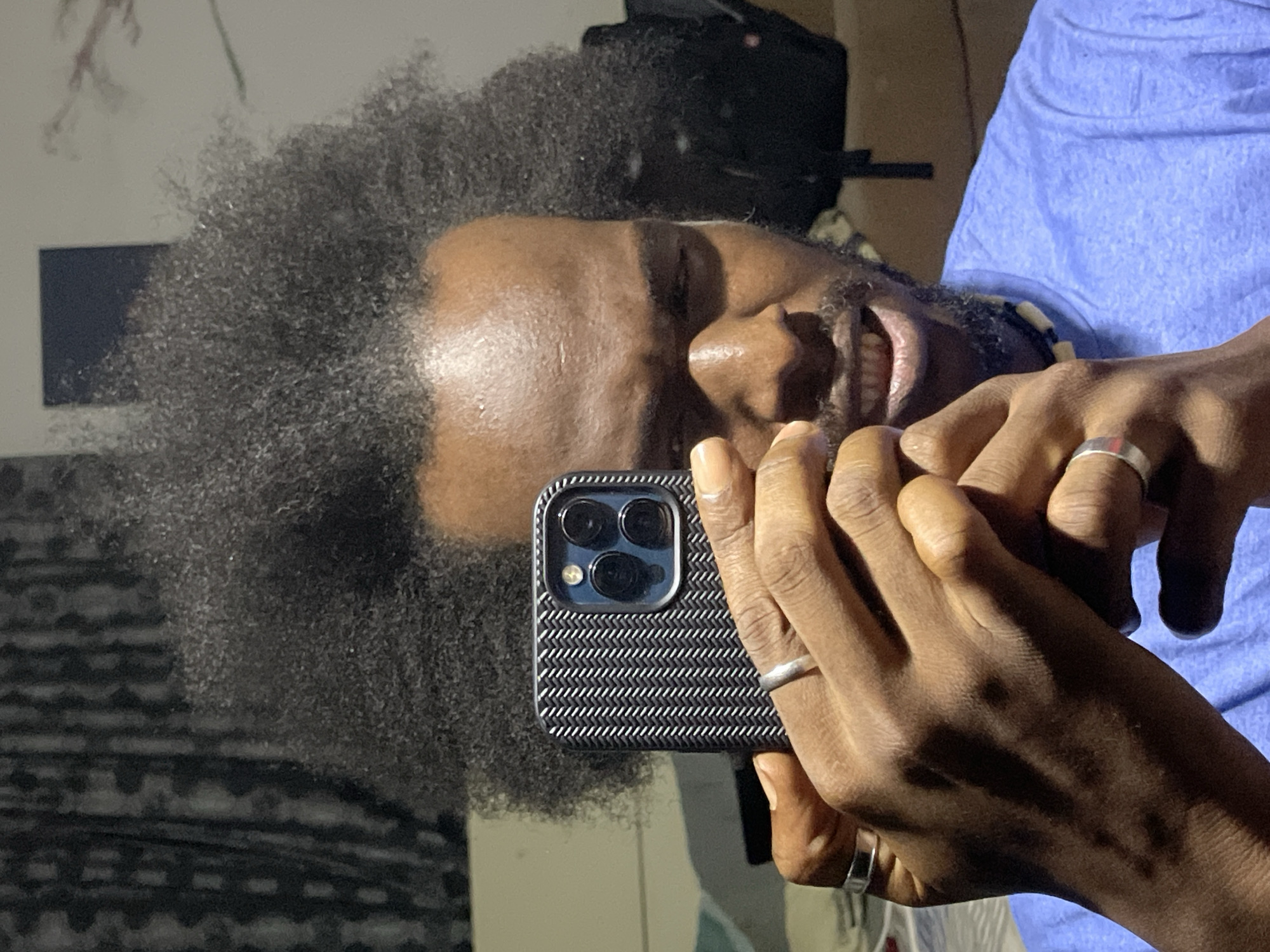 A man with dark skin and a large Afro hairstyle is holding a smartphone with three camera lenses in front of his face. The man is smiling, revealing his teeth. He is wearing a light blue shirt. He has multiple rings on his fingers. In the background, there is a dark backpack and a patterned wall. The location is not discernible as Kuchingoro, Nigeria.